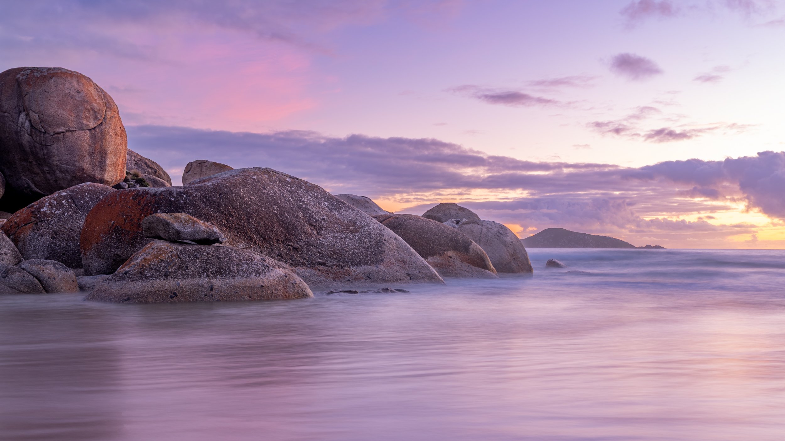 Scenic view of large rocks along a calm beach at sunset with pastel-colored sky and distant island.