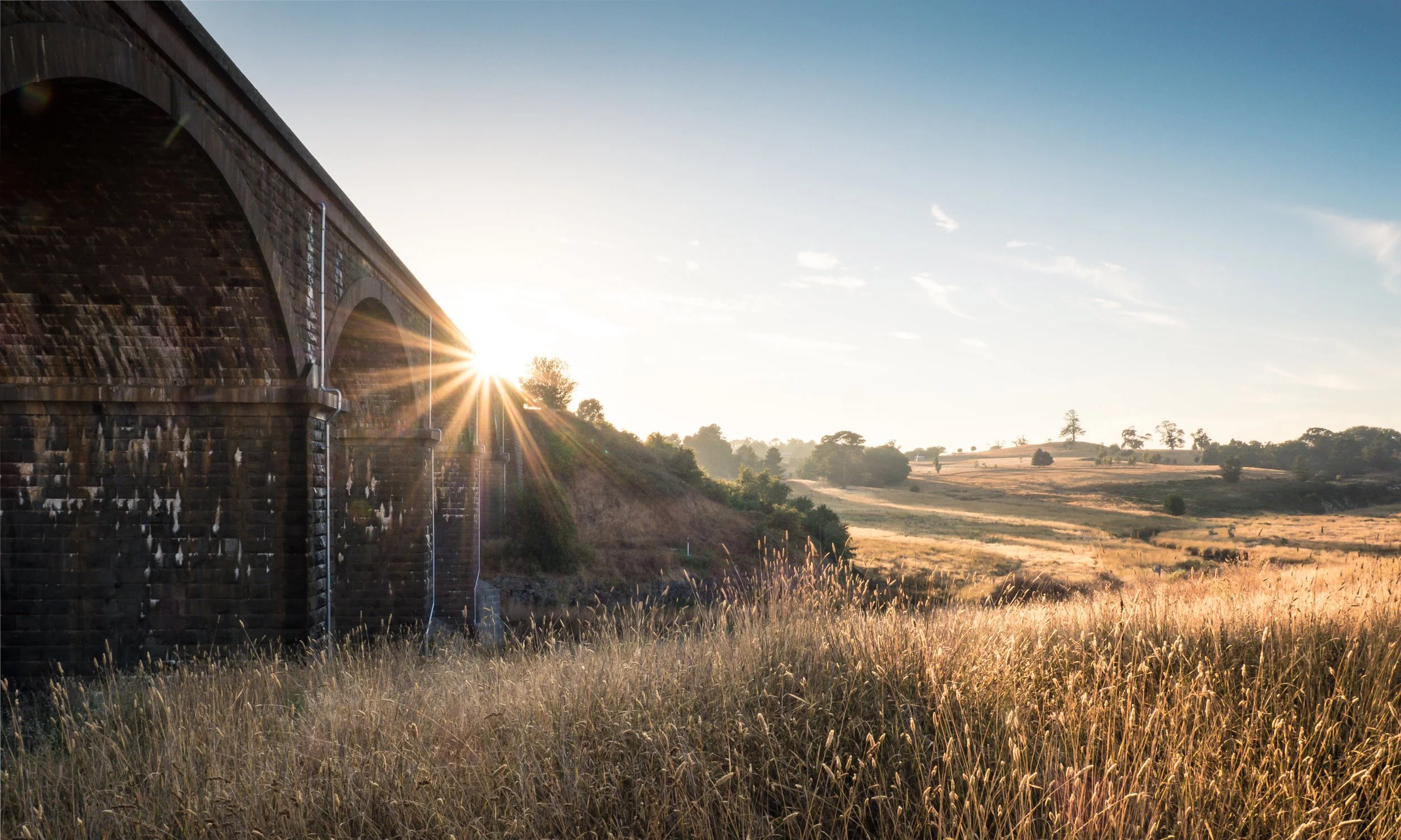 Sunset over a rural landscape with an old stone bridge on the left, tall grasses in the foreground, rolling hills and trees in the distance, and a clear blue sky.