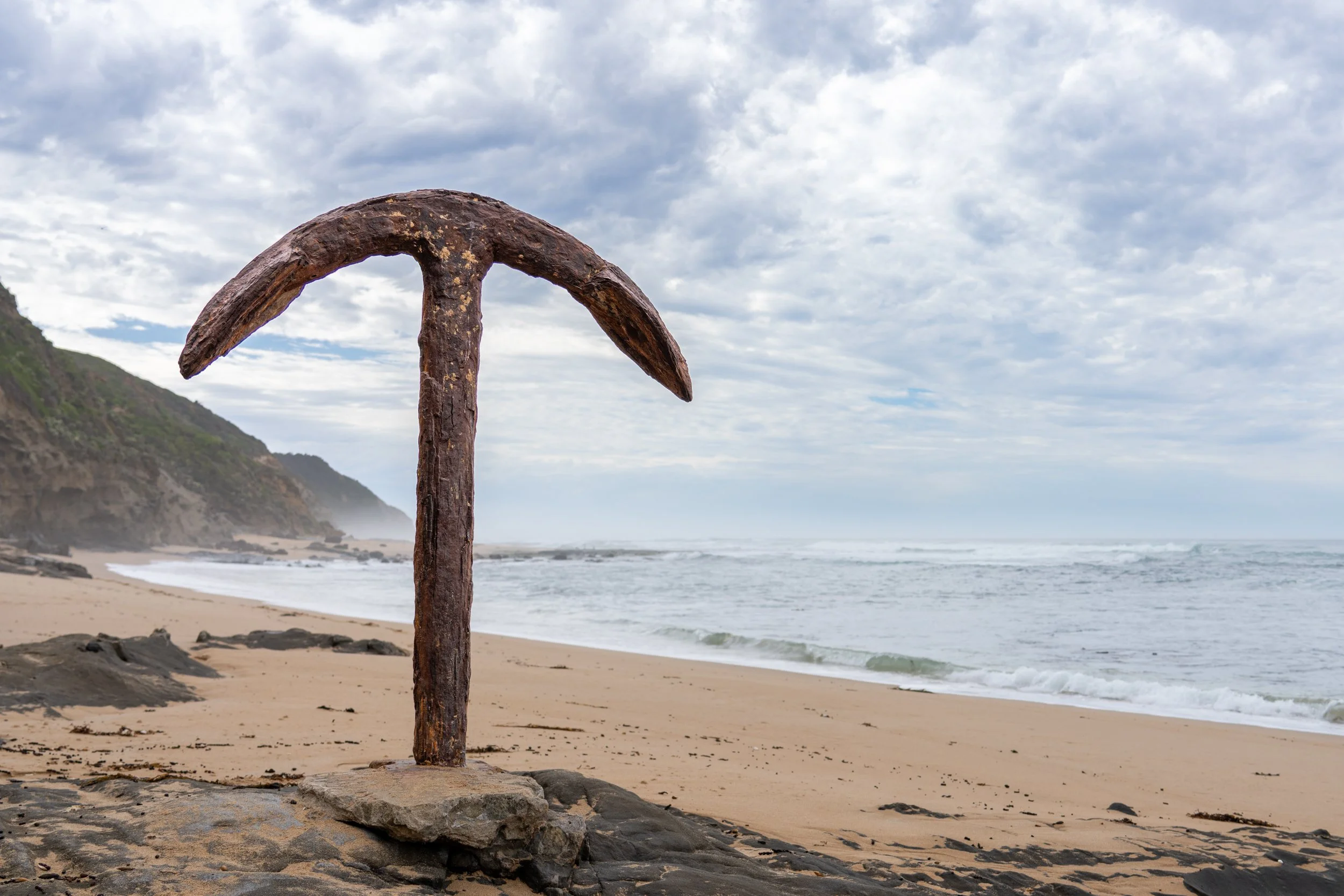 A rusted ship anchor on the sandy beach with ocean waves and cloudy sky in the background.