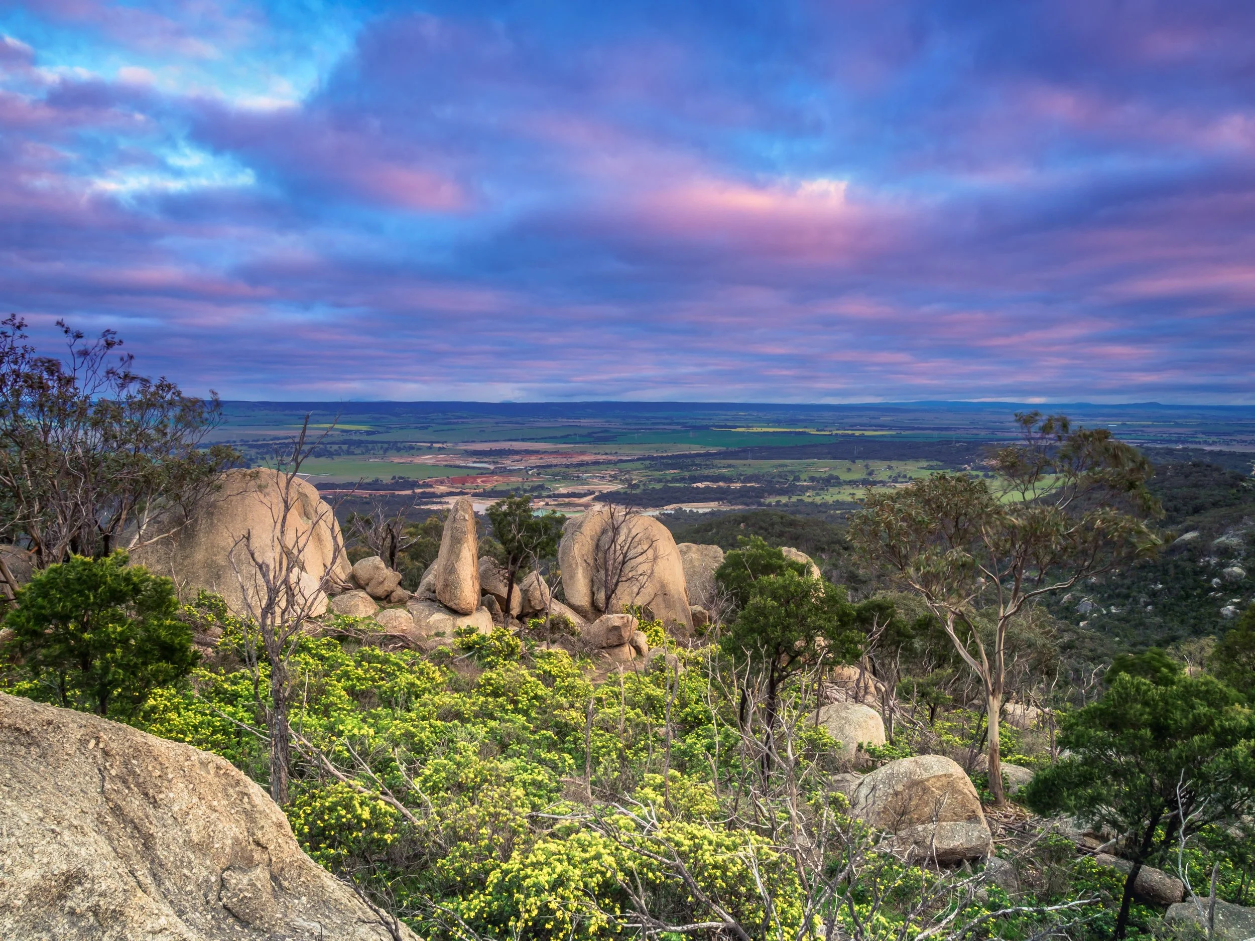 A scenic landscape with large boulders and green shrubbery in the foreground, rolling hills and farmland in the background, and a colorful, cloudy sky above.