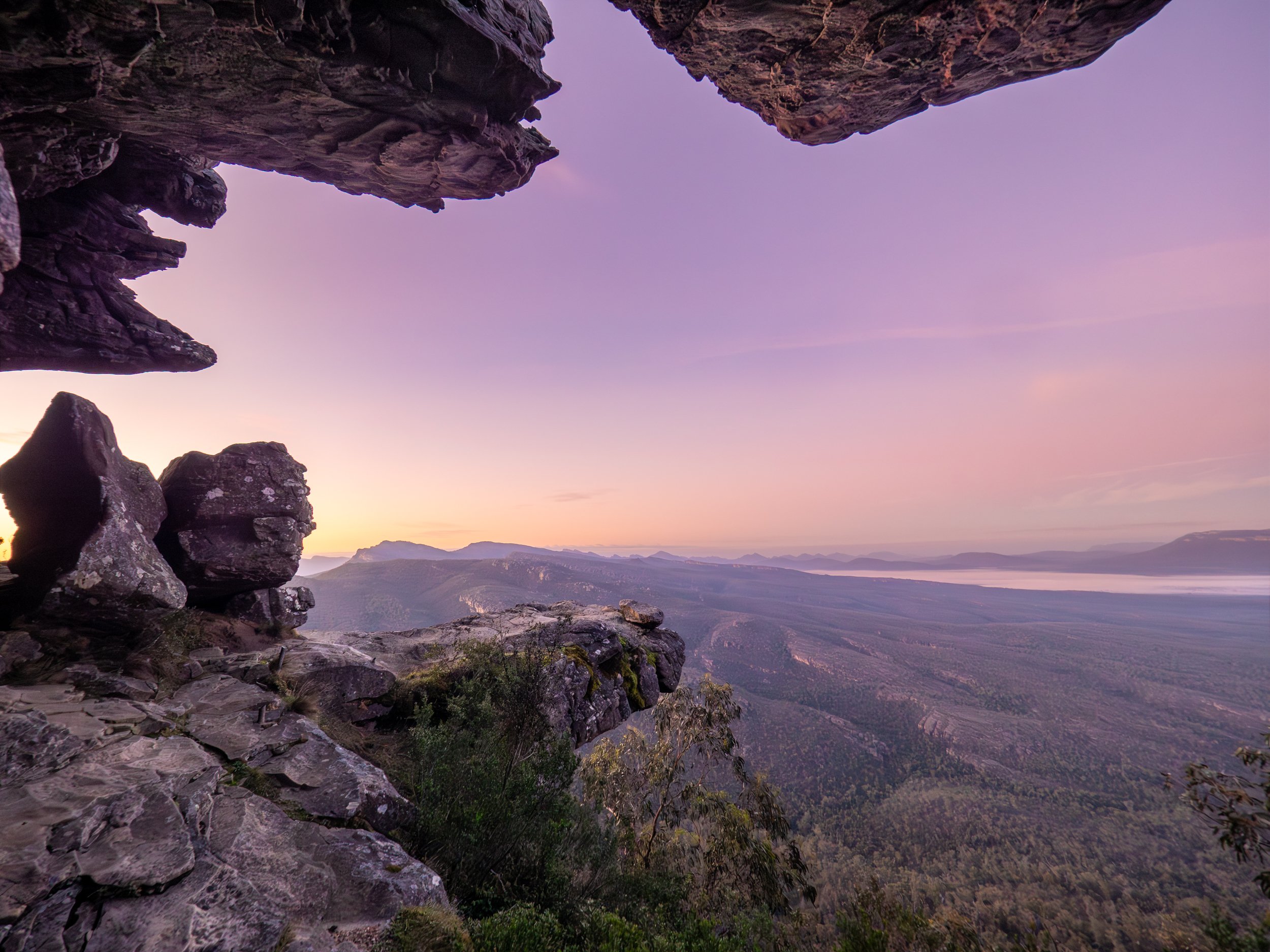 View from a rocky cliff overlooking a vast valley at sunset with pink and purple sky.