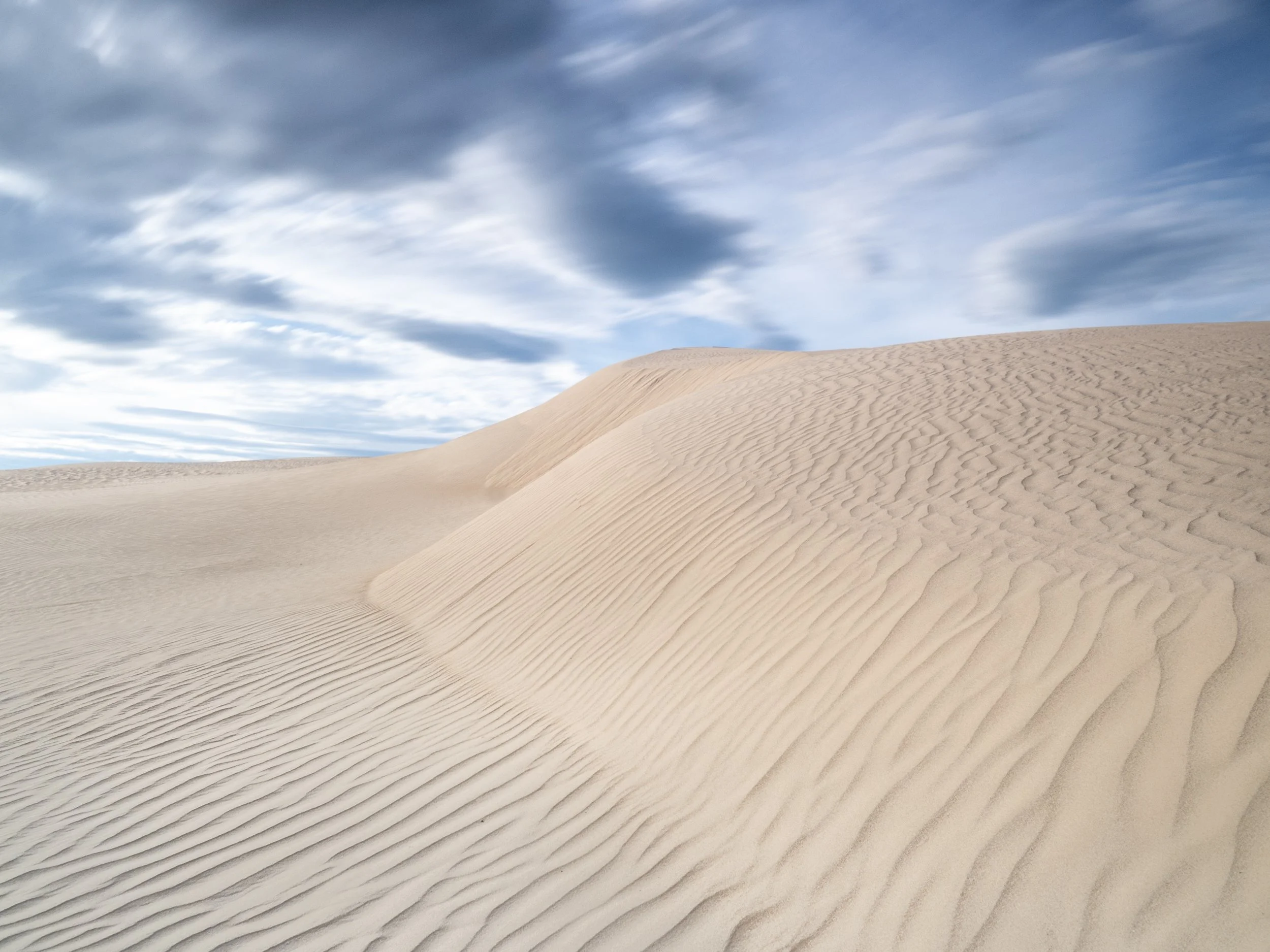 Sand dunes under a partly cloudy sky with streaks of clouds.