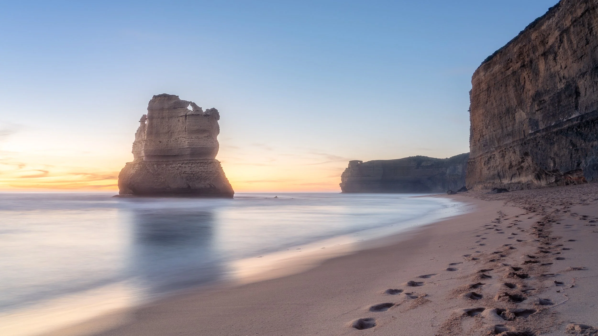 Beach at sunset with large rock formations and footprints in the sand.