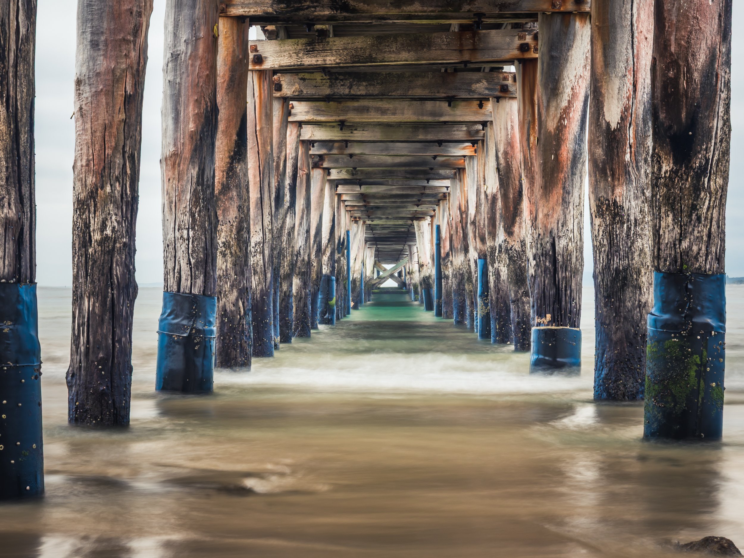 Underneath a wooden pier extending into the ocean, with weathered wooden beams and supports, some covered in black protective sheeting and green moss, with water flowing around the supports.
