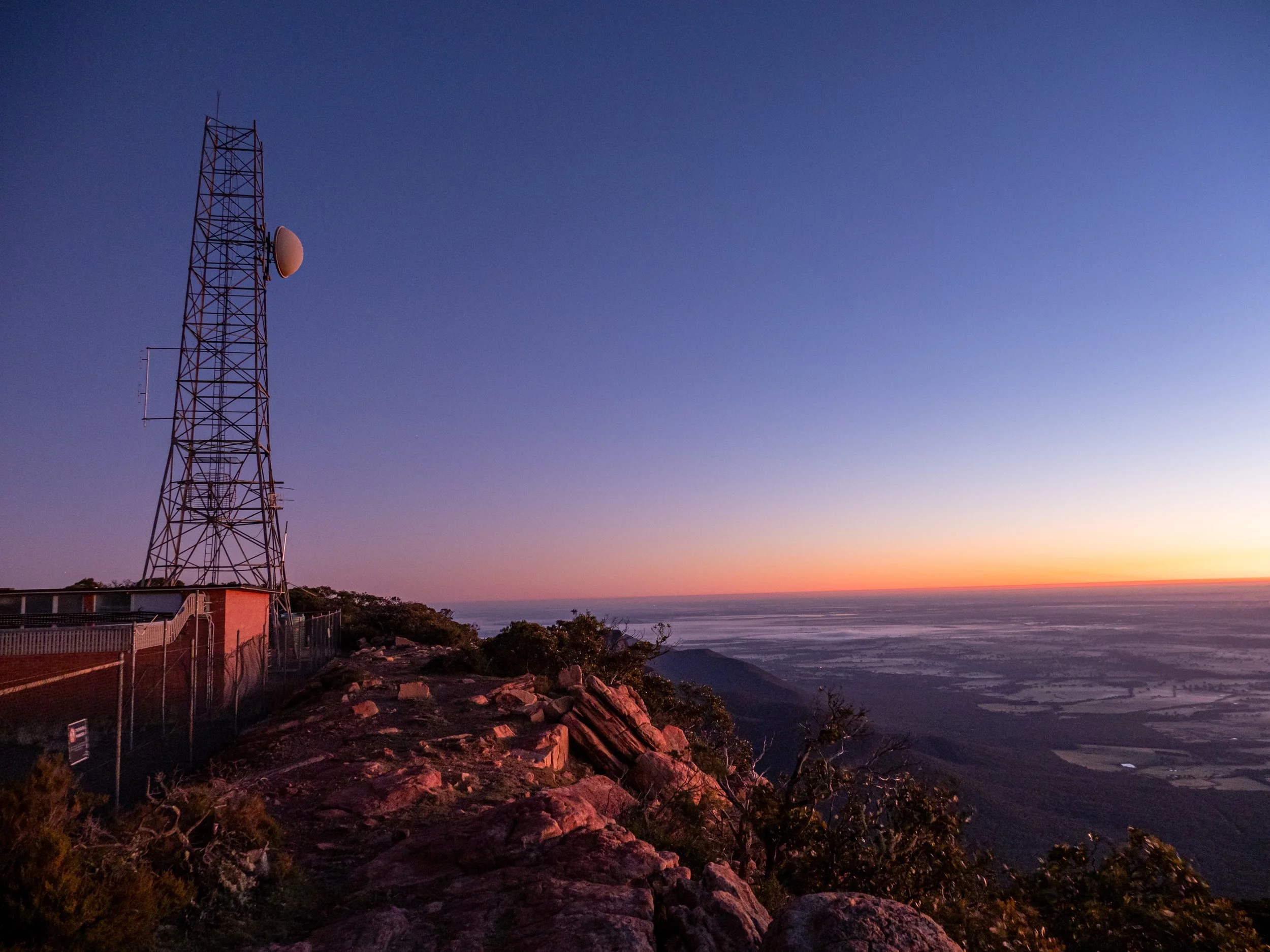 A tall communication tower on a hilltop with a fence, overlooking a landscape during sunset with a colorful sky.
