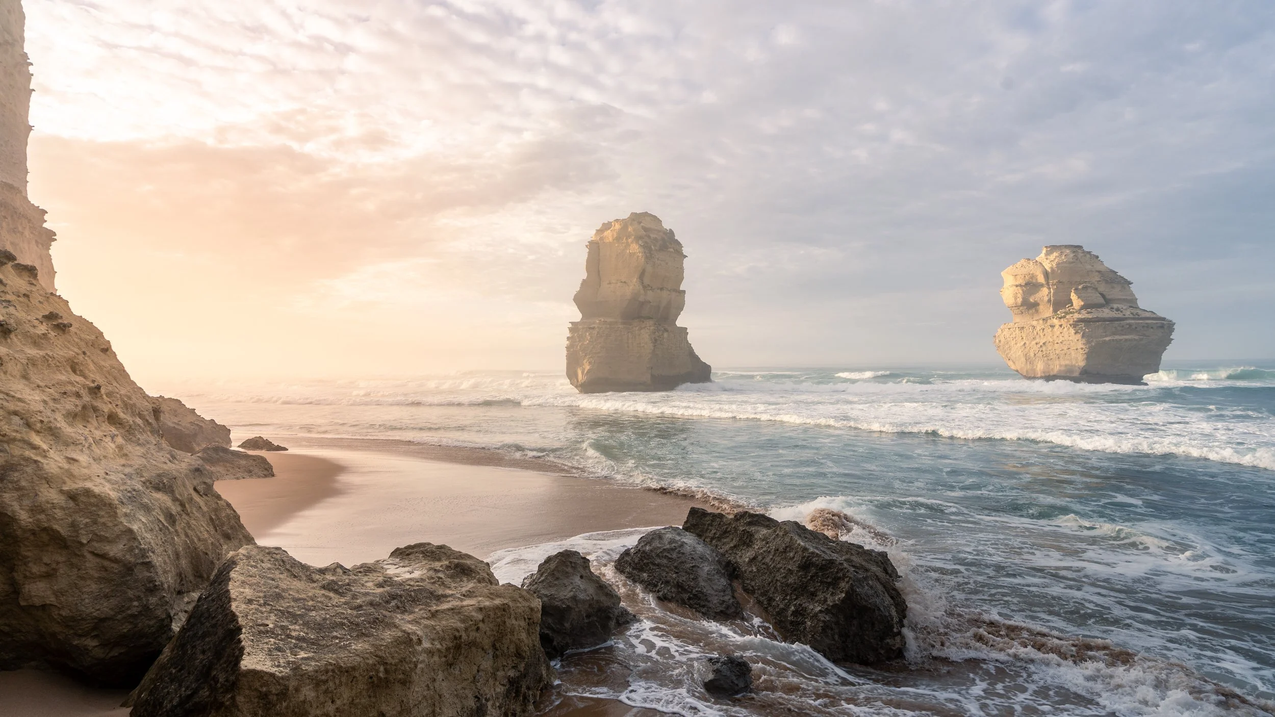 A coastal scene with large rocks and sandy beach in the foreground, and two large sea stacks in the ocean under a partly cloudy sky, with soft sunlight illuminating the landscape.