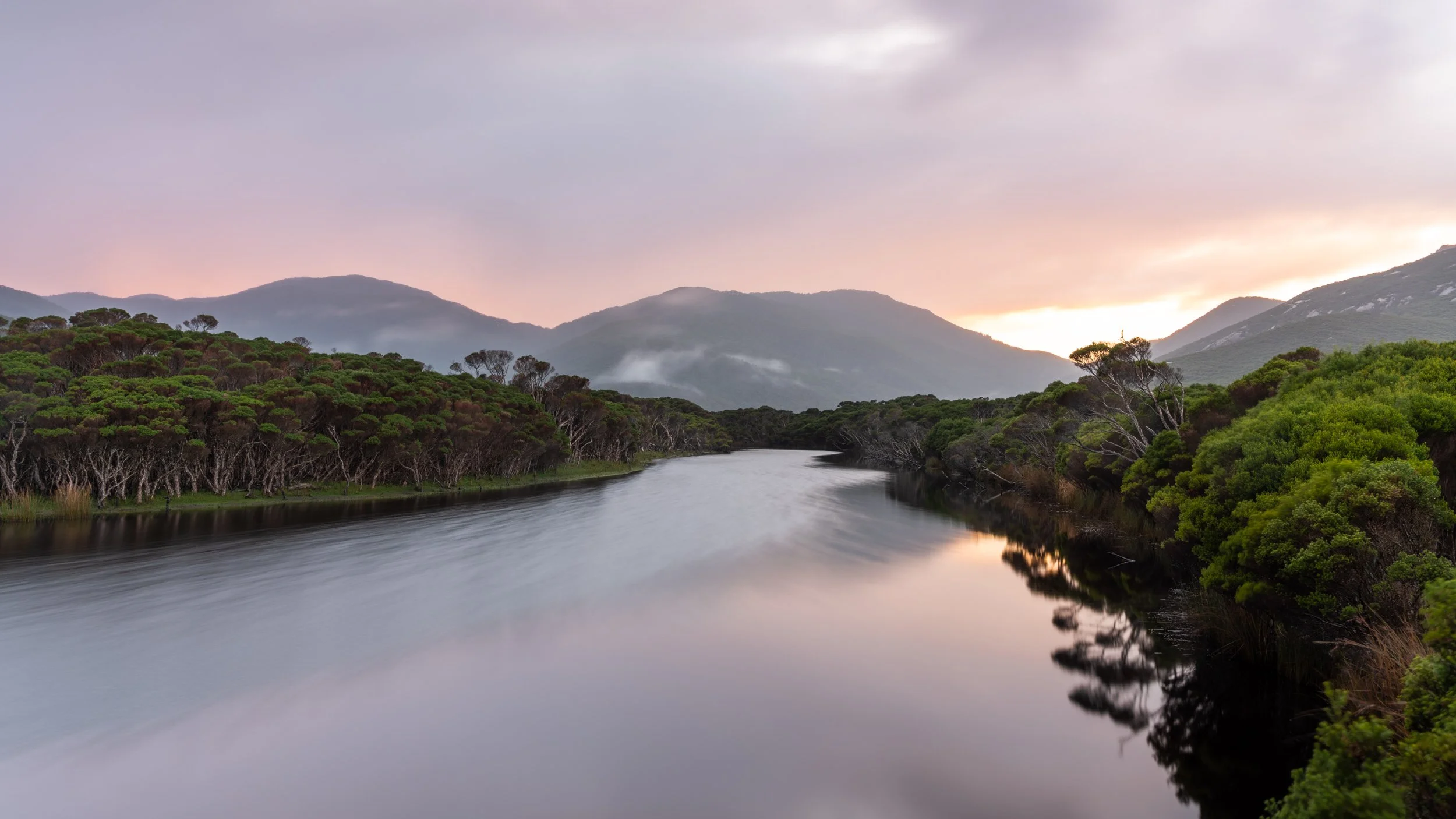 A calm river flows through a lush green landscape with trees on both sides, mountains in the background, and a pastel-colored sky at sunset.