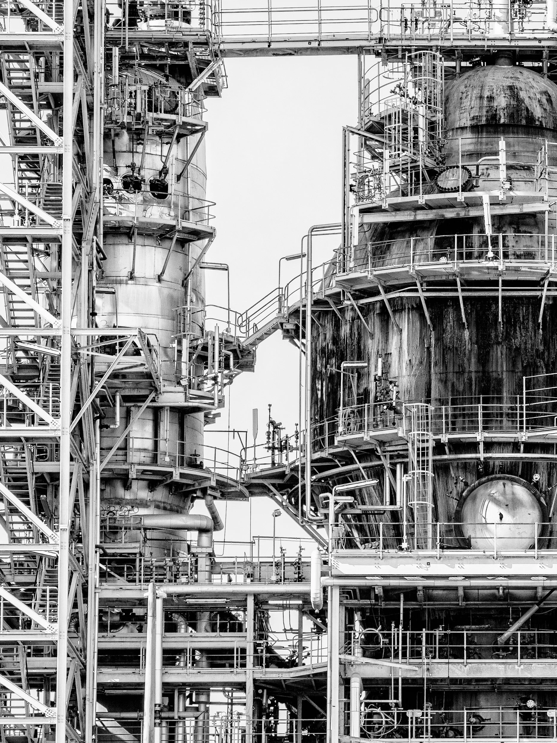 Black and white image of an industrial refinery with large cylindrical tanks, pipes, and metal staircases.