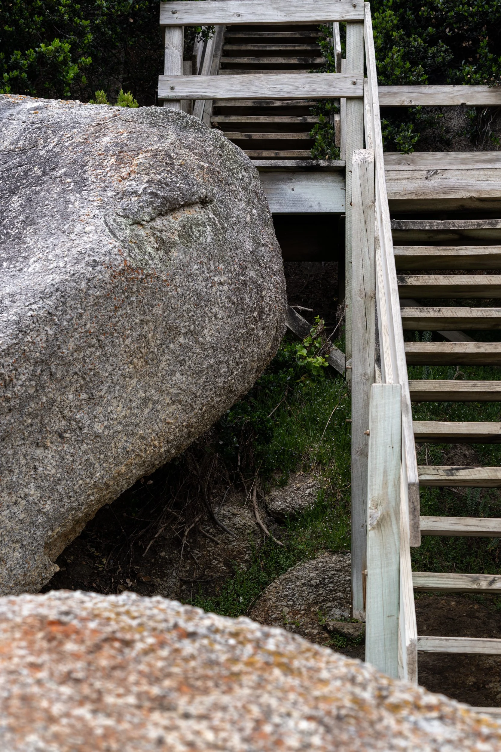A large boulder next to a wooden staircase outdoors, with green bushes in the background.