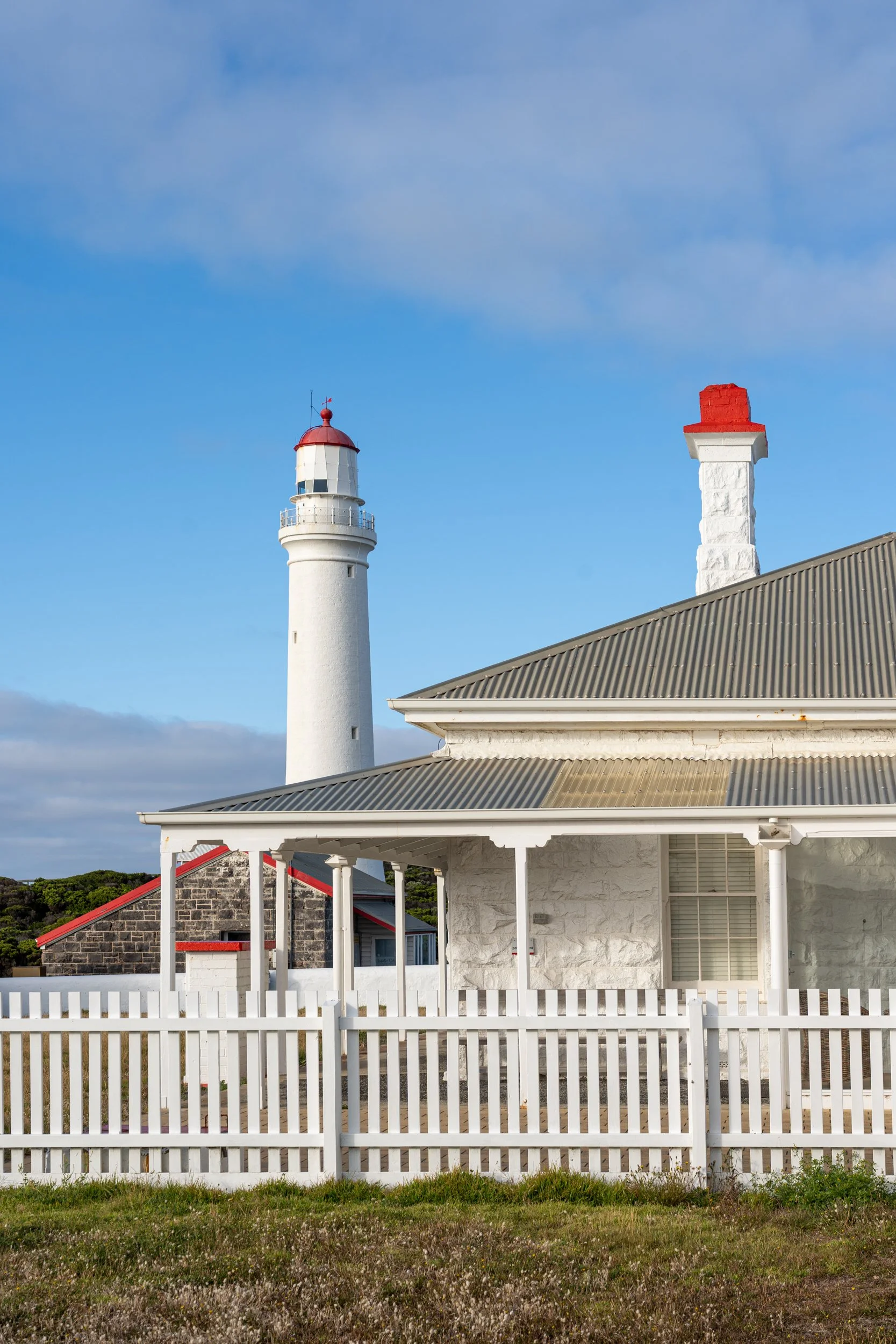 A white lighthouse with a red top next to a white house with a grey metal roof and white fence, under a partly cloudy blue sky.
