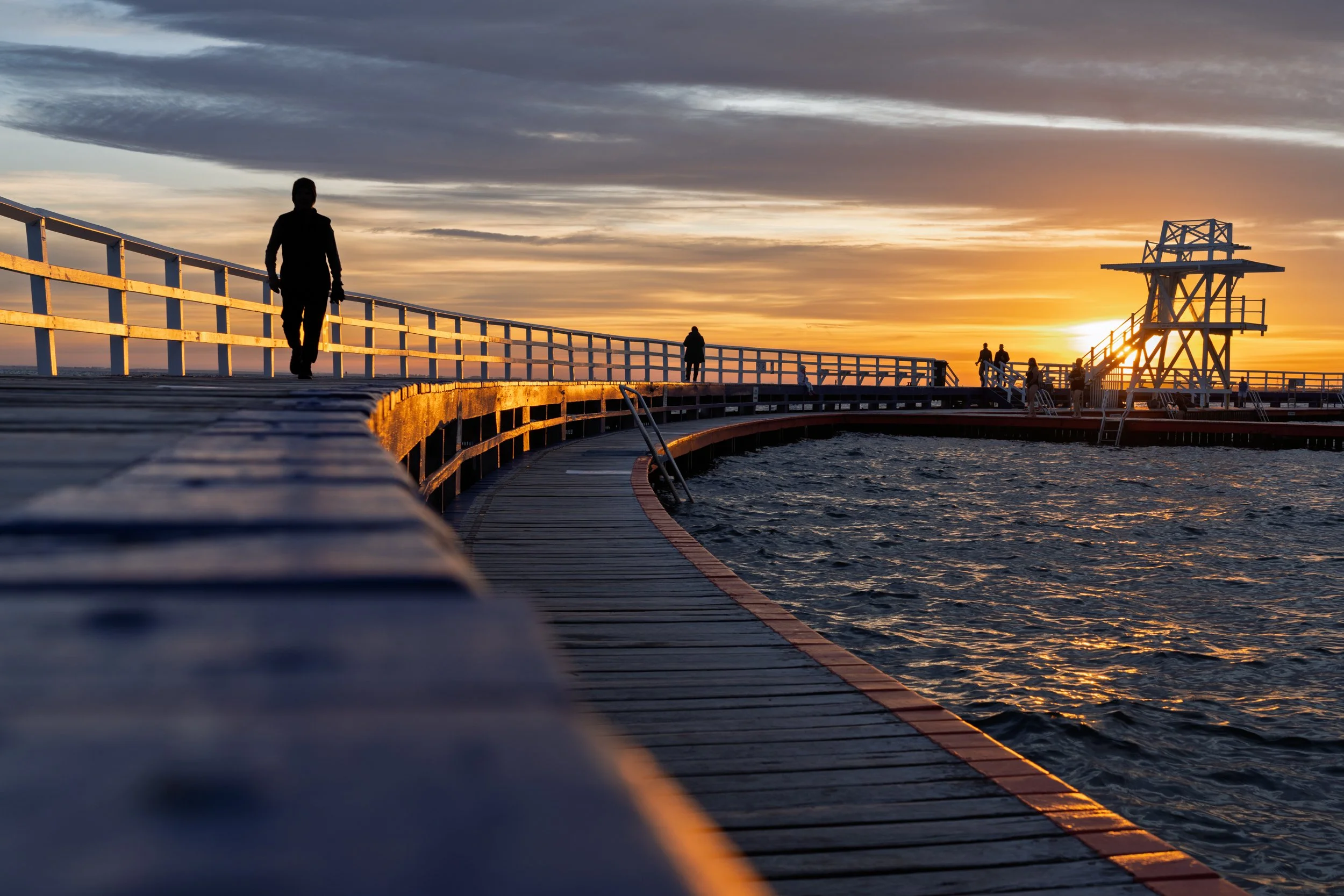Silhouettes of people walking on a curved wooden pier during sunset, with a lifeguard tower and the ocean visible.