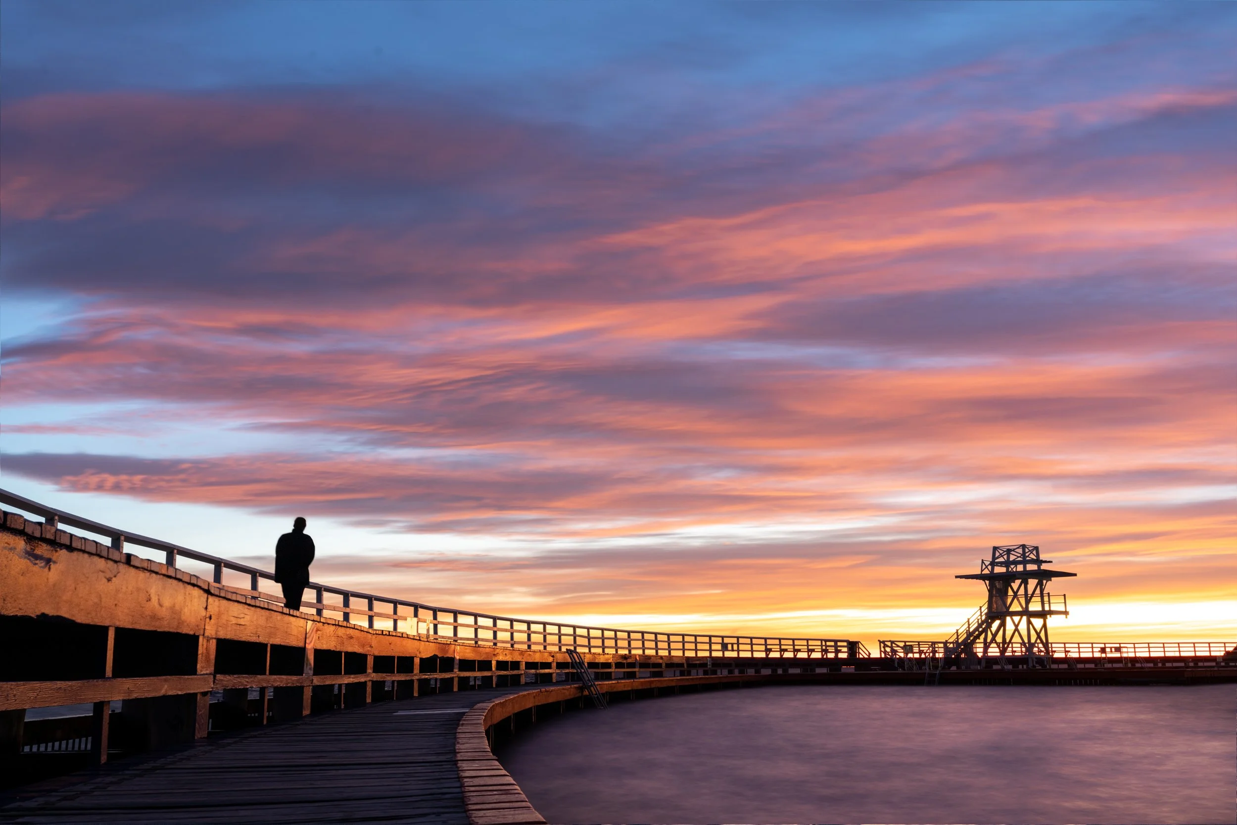 A silhouette of a man walking on a curved wooden pier at sunset with a colorful sky and a lighthouse structure in the distance.