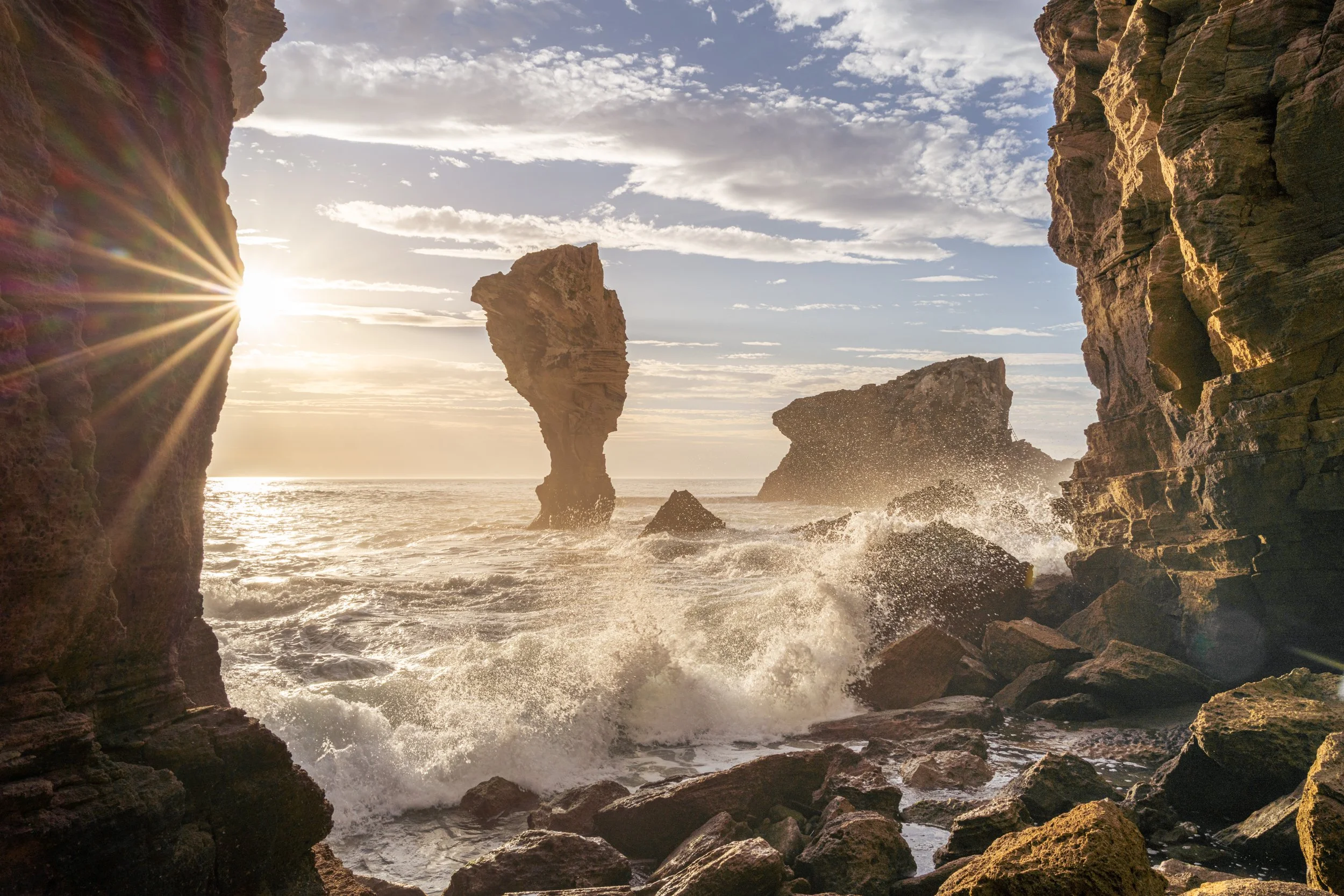 Sunset over rocky coastal cliffs with waves crashing and large sea stacks in the ocean.