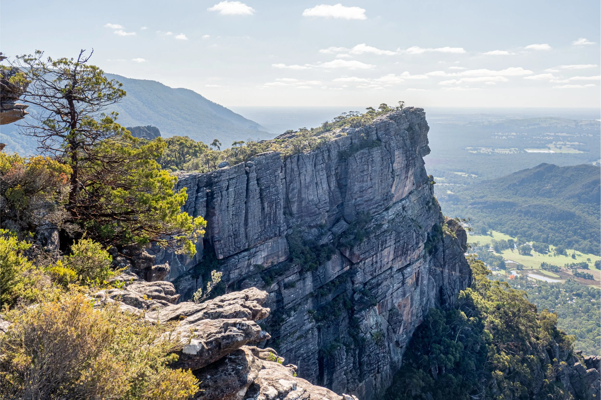 A scenic view of a rocky cliff with lush greenery, overlooking a forested valley and distant mountains under a partly cloudy sky.