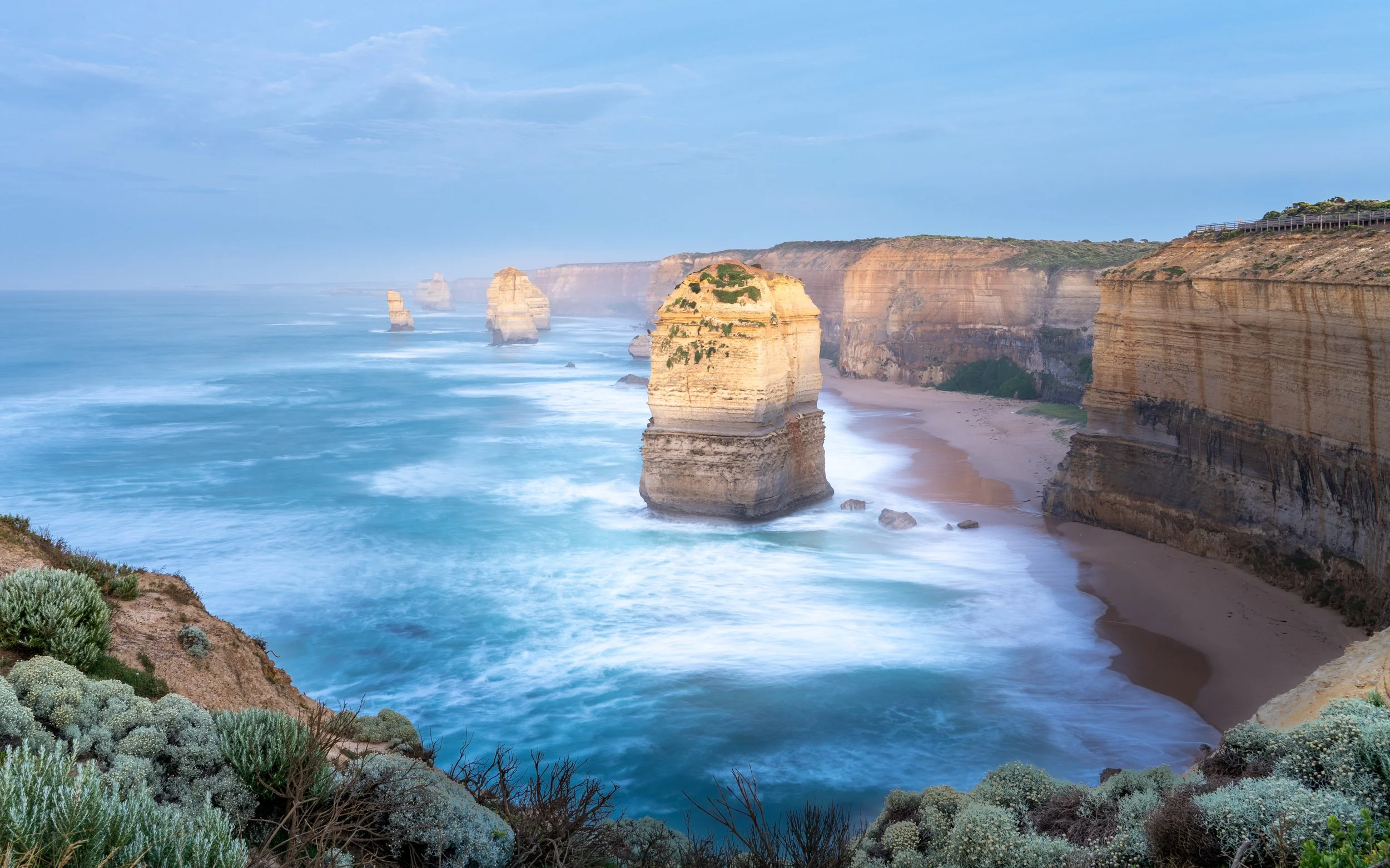 Coastal view of the Twelve Apostles limestone stacks along the Great Ocean Road in Australia, with rugged cliffs, sandy beach, and lush vegetation in the foreground.