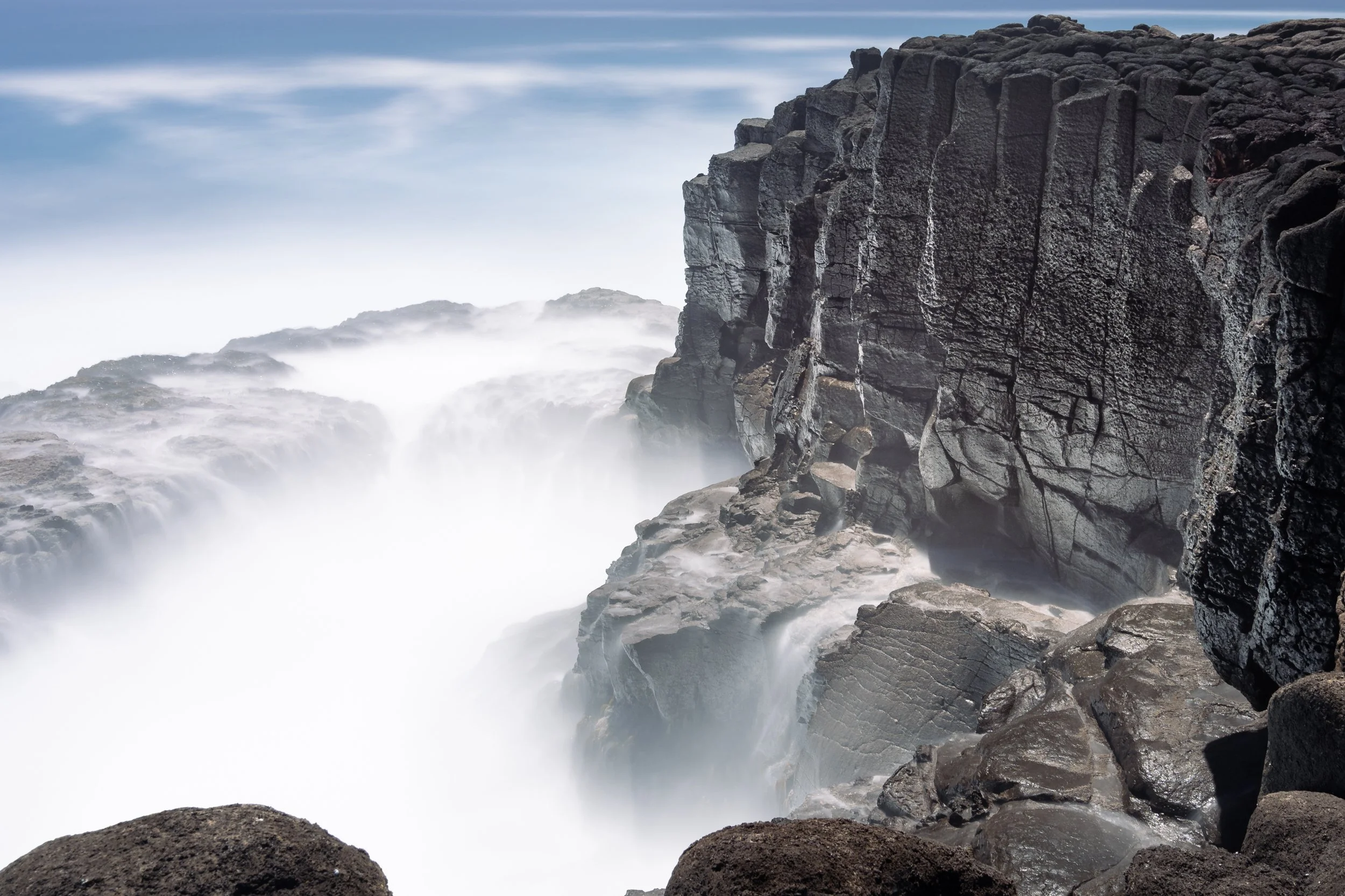 Coastal scene with crashing ocean waves hitting dark rocky cliffs, mist rising from the water, and a partly cloudy sky above.