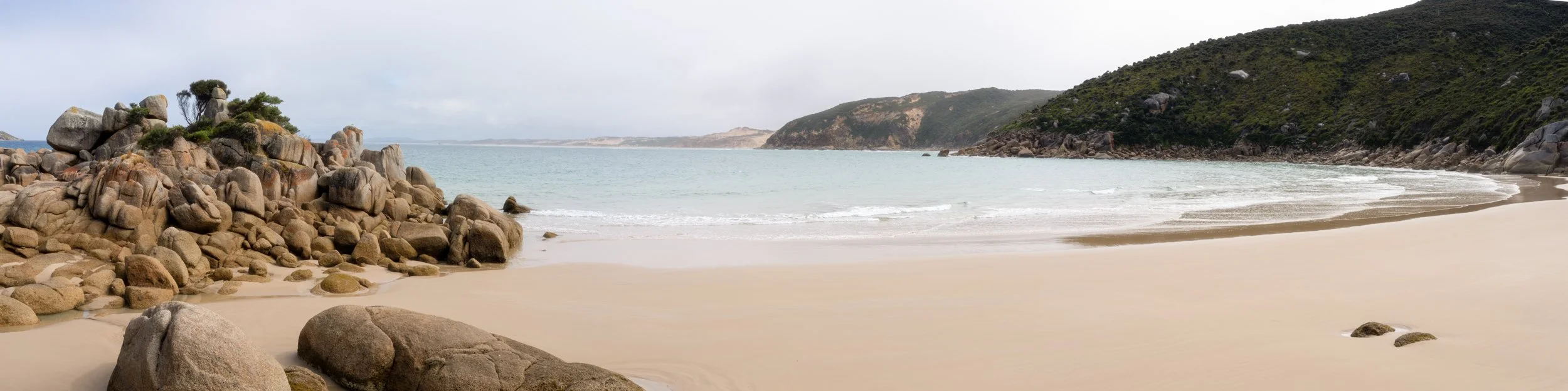 A beach with rocks on the left, sandy shore in the foreground, ocean in the middle, and hills covered with greenery in the background.