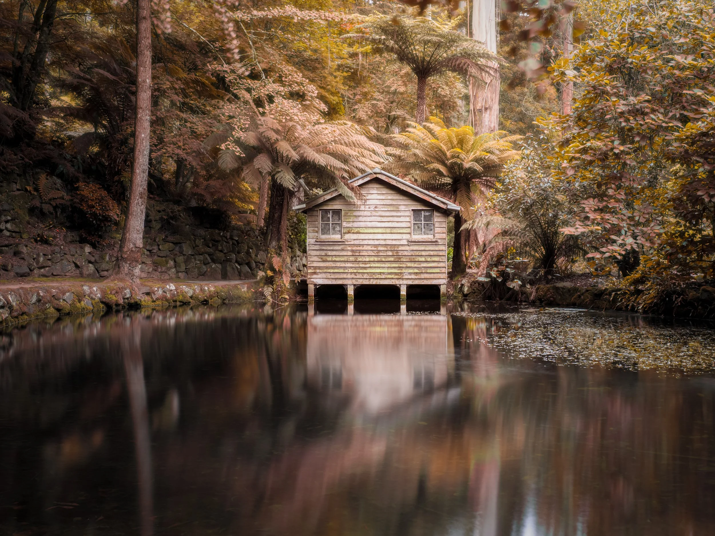 A rustic wooden boathouse on stilts by a calm river, surrounded by lush trees with autumn leaves, in a peaceful forest scene.