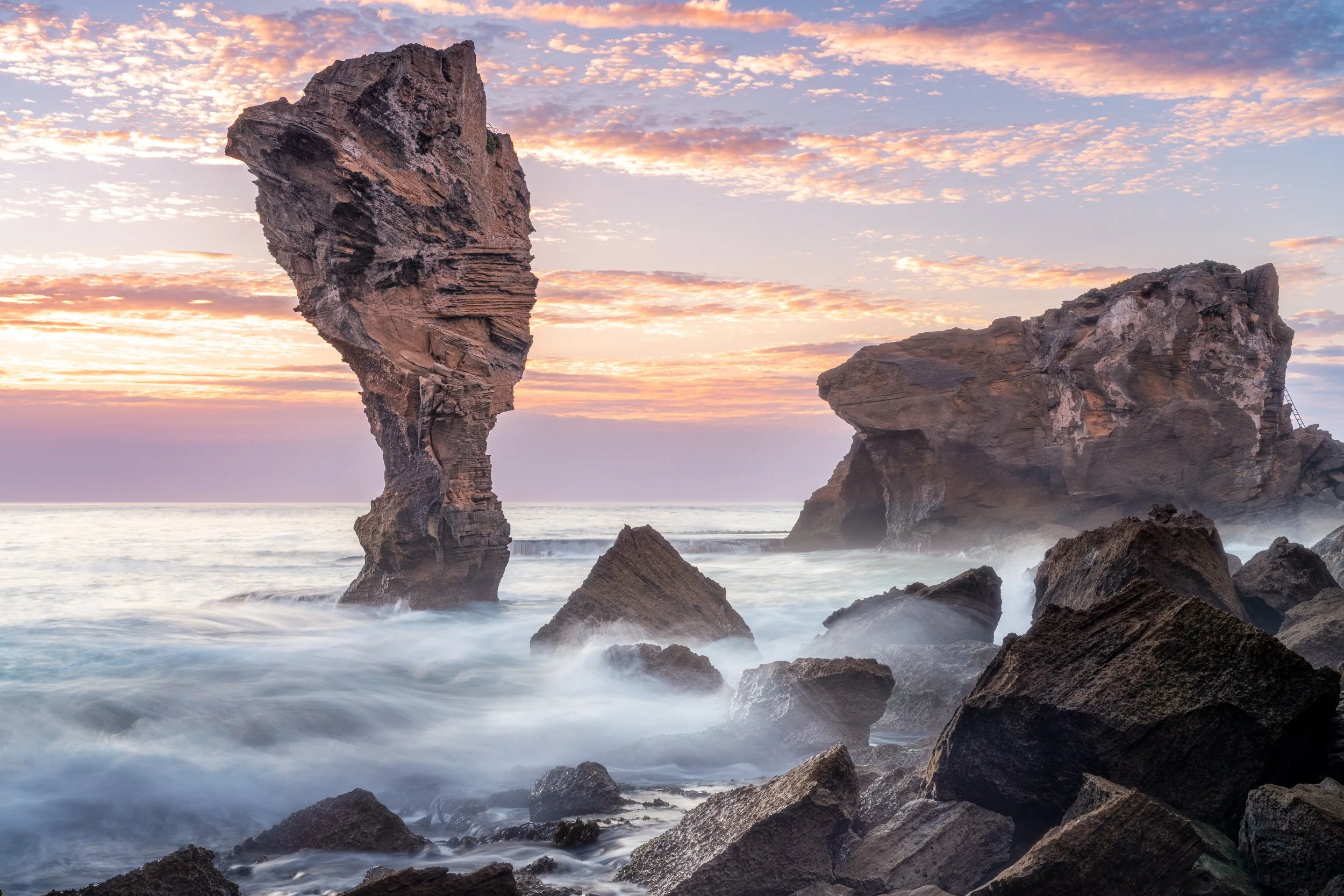 Sunset over rugged coastal rocks and formations, with a distinctive tall rock formation on the left and a large rock on the right, ocean waves crashing against the rocks, and a colorful sky with scattered clouds.
