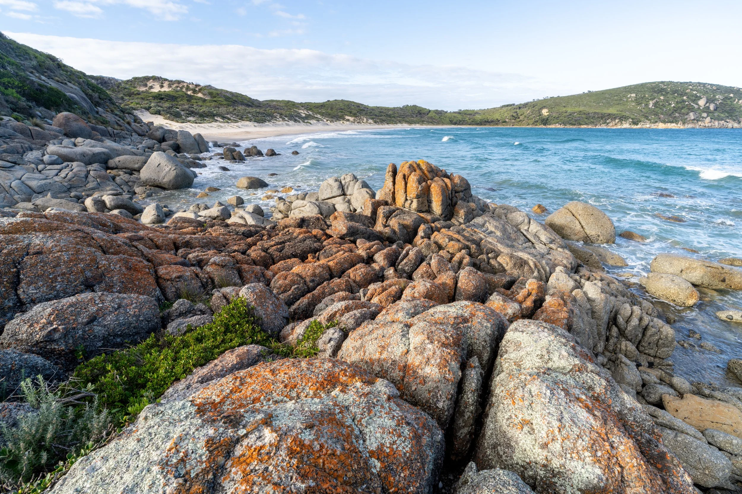 Rocky shoreline with ocean waves and distant green hills under partly cloudy sky.
