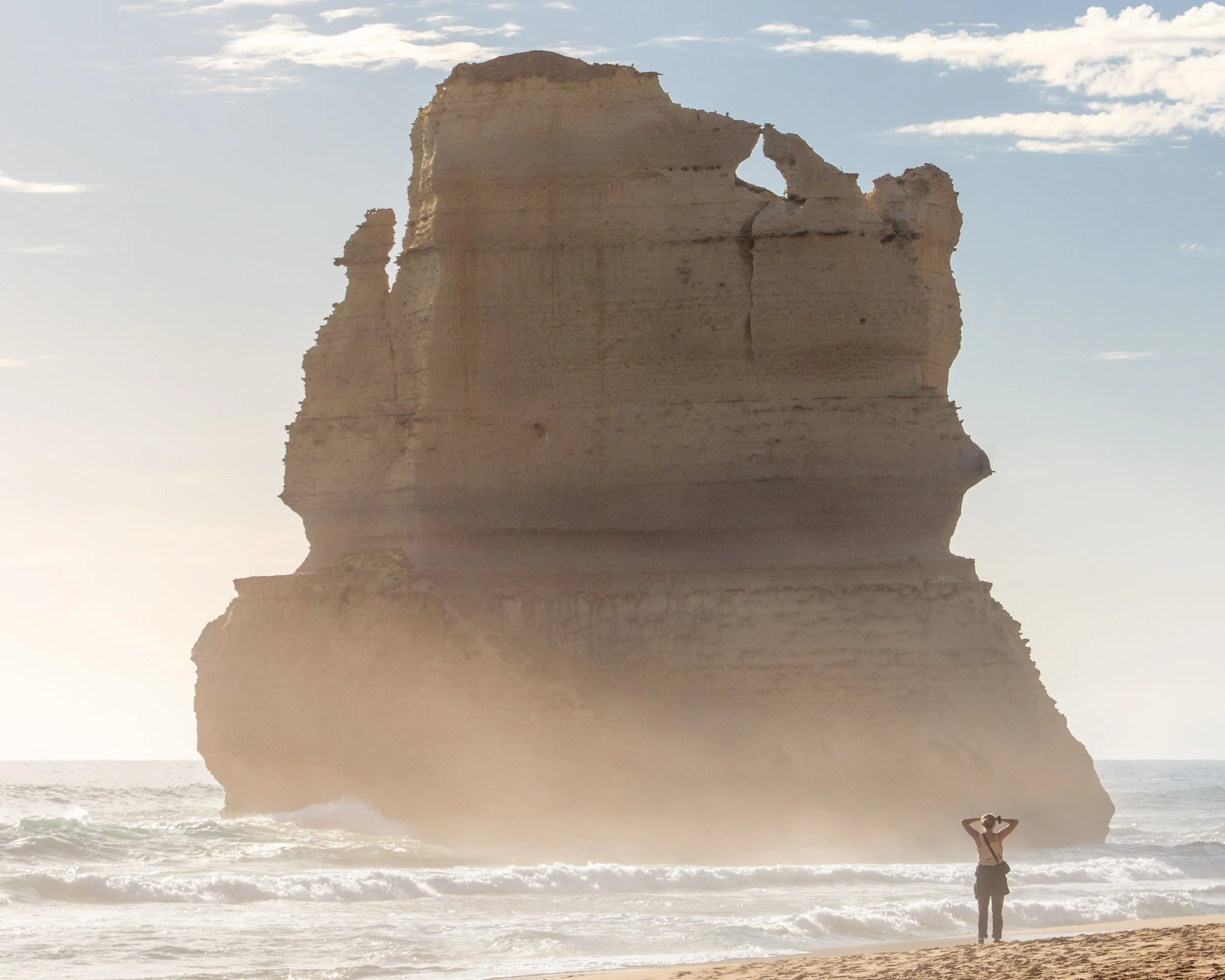 A person standing on a sandy beach, looking at a large sea stack rock formation in the ocean with waves crashing around its base, under a partly cloudy sky.