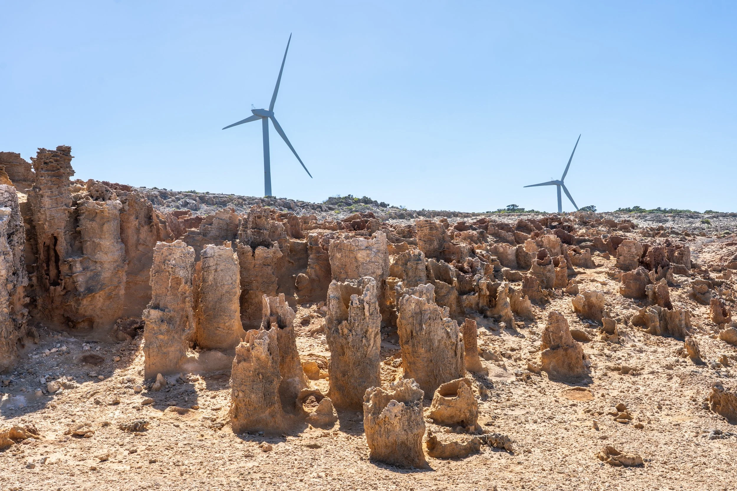 A landscape with rocky terrain and formations, two wind turbines in the background under a clear blue sky.