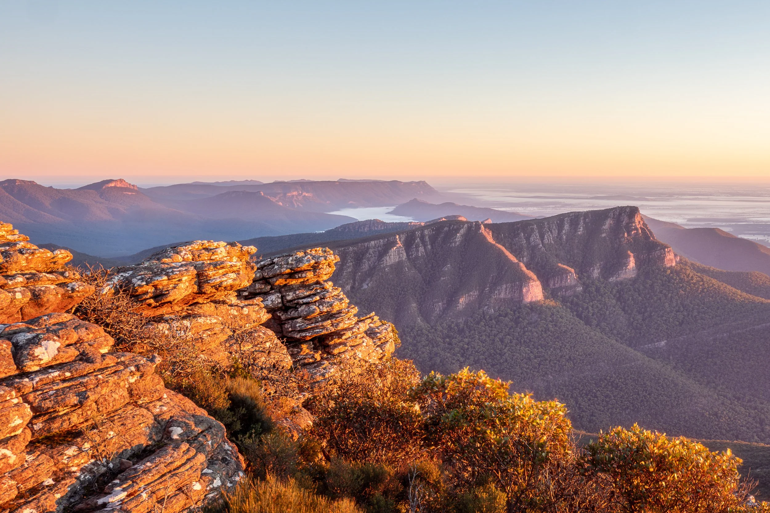 Sunrise over mountain range with layered hills, rocky foreground, and sparse vegetation.
