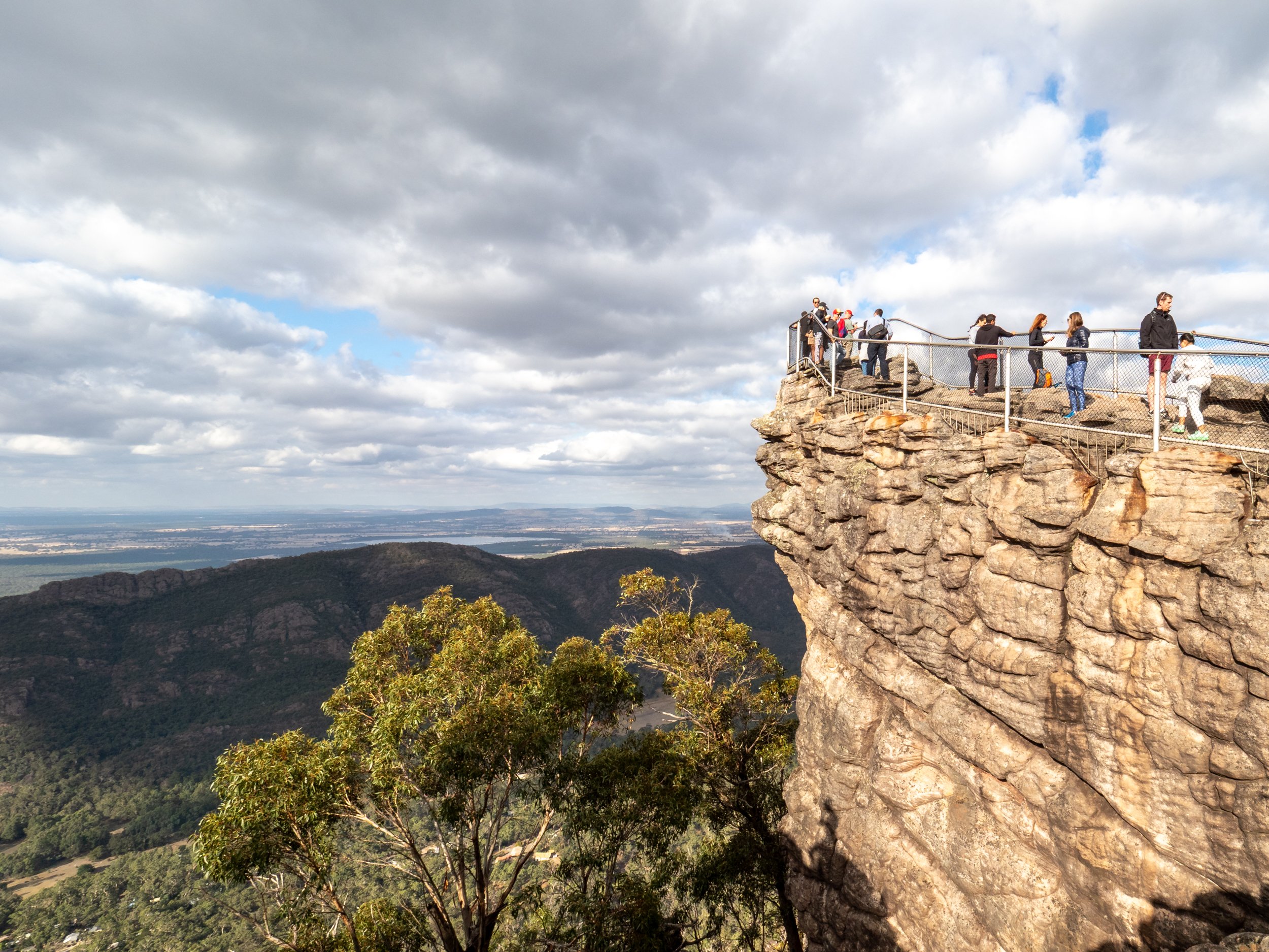 People standing on a viewing platform at the edge of a rocky cliff, overlooking a valley with mountains in the distance under a partly cloudy sky.