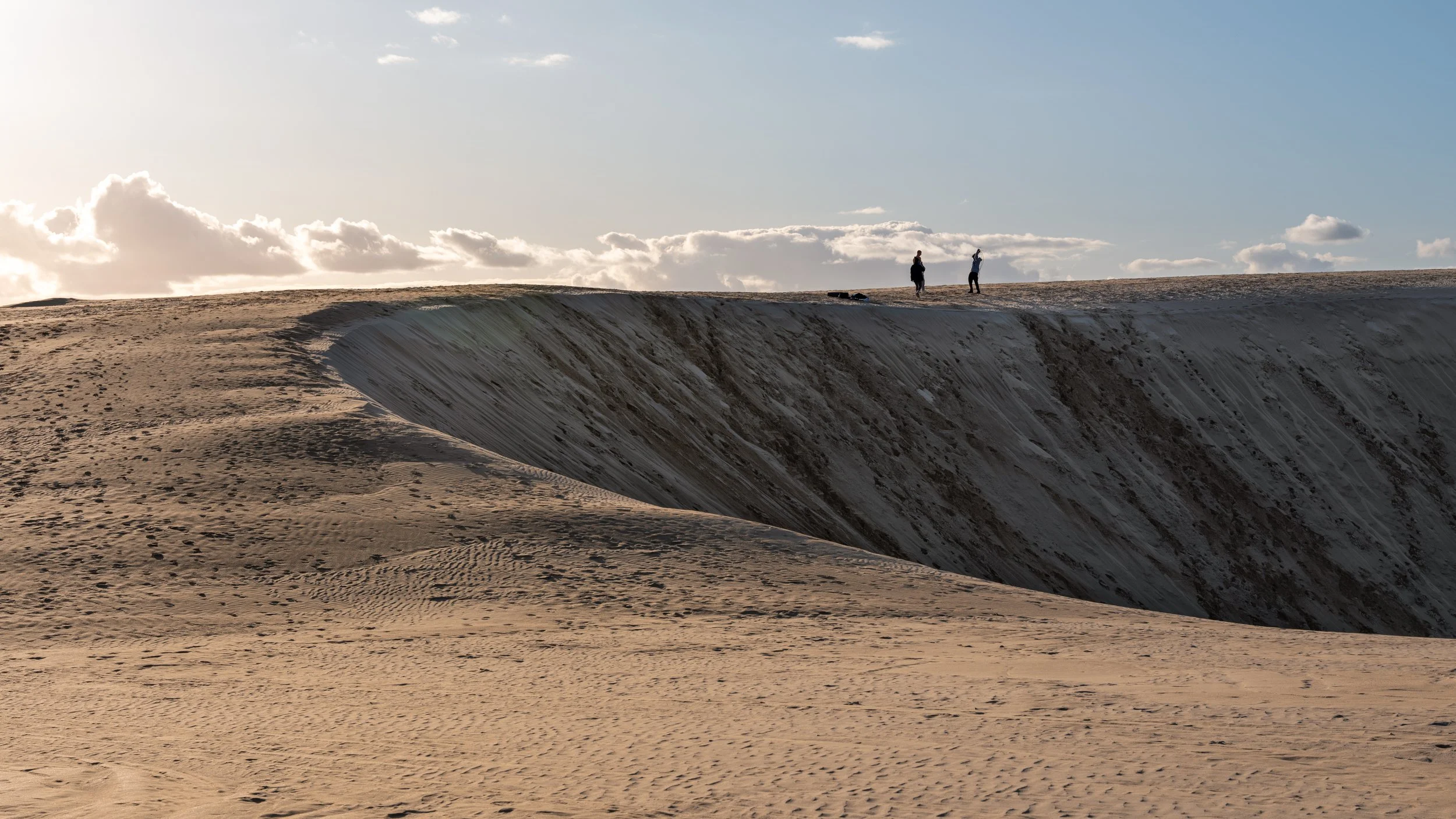 Two people standing on the edge of a large sand dune in a desert, with clouds and a blue sky in the background.