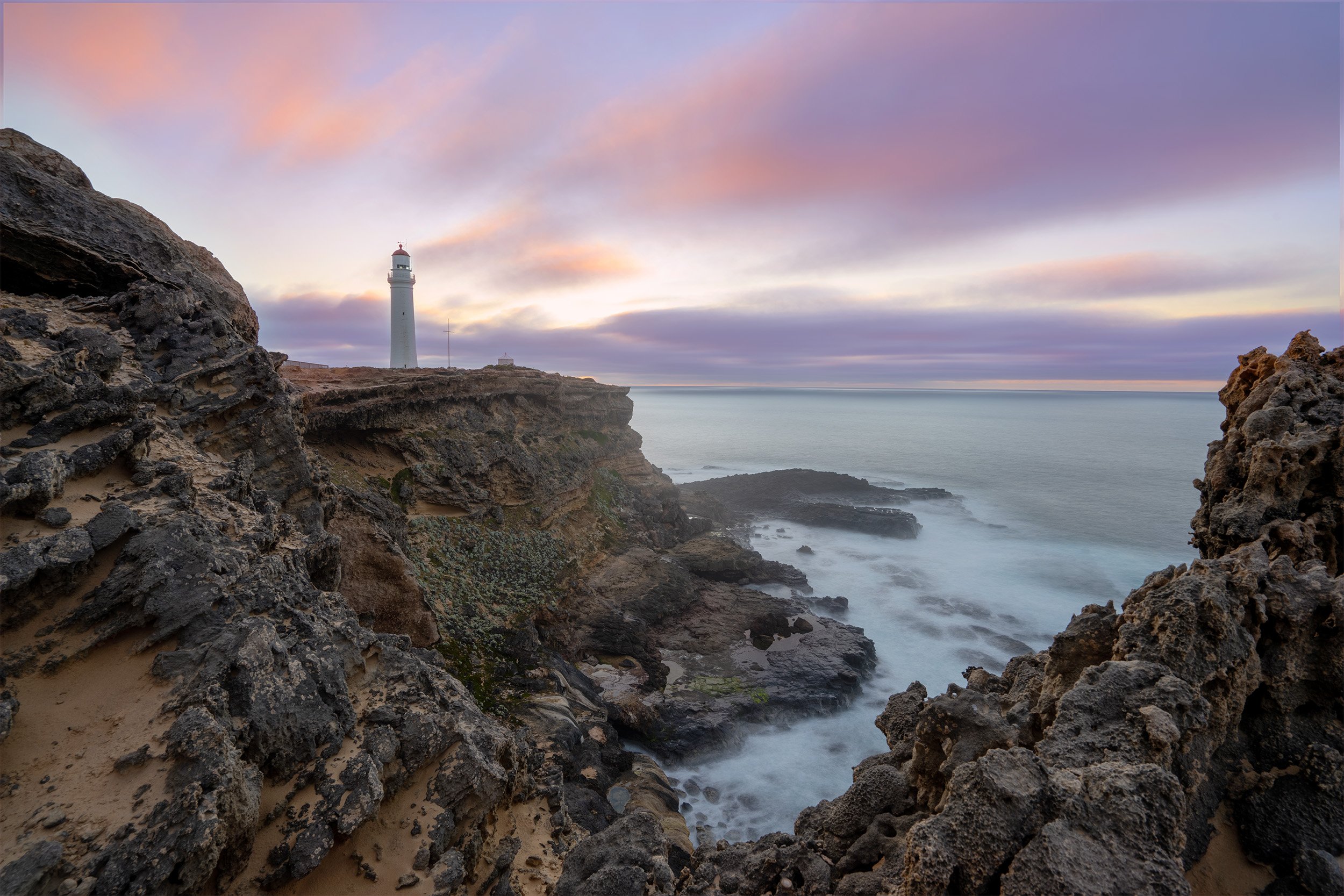 A lighthouse on a cliff overlooking the ocean during a colorful sunset with purple and pink clouds.