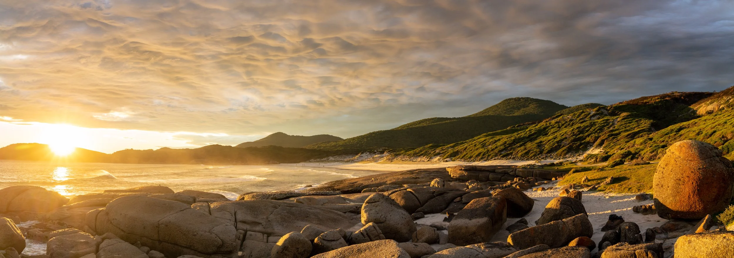 Sun setting over a rocky beach with hills covered in greenery in the background and a partly cloudy sky.