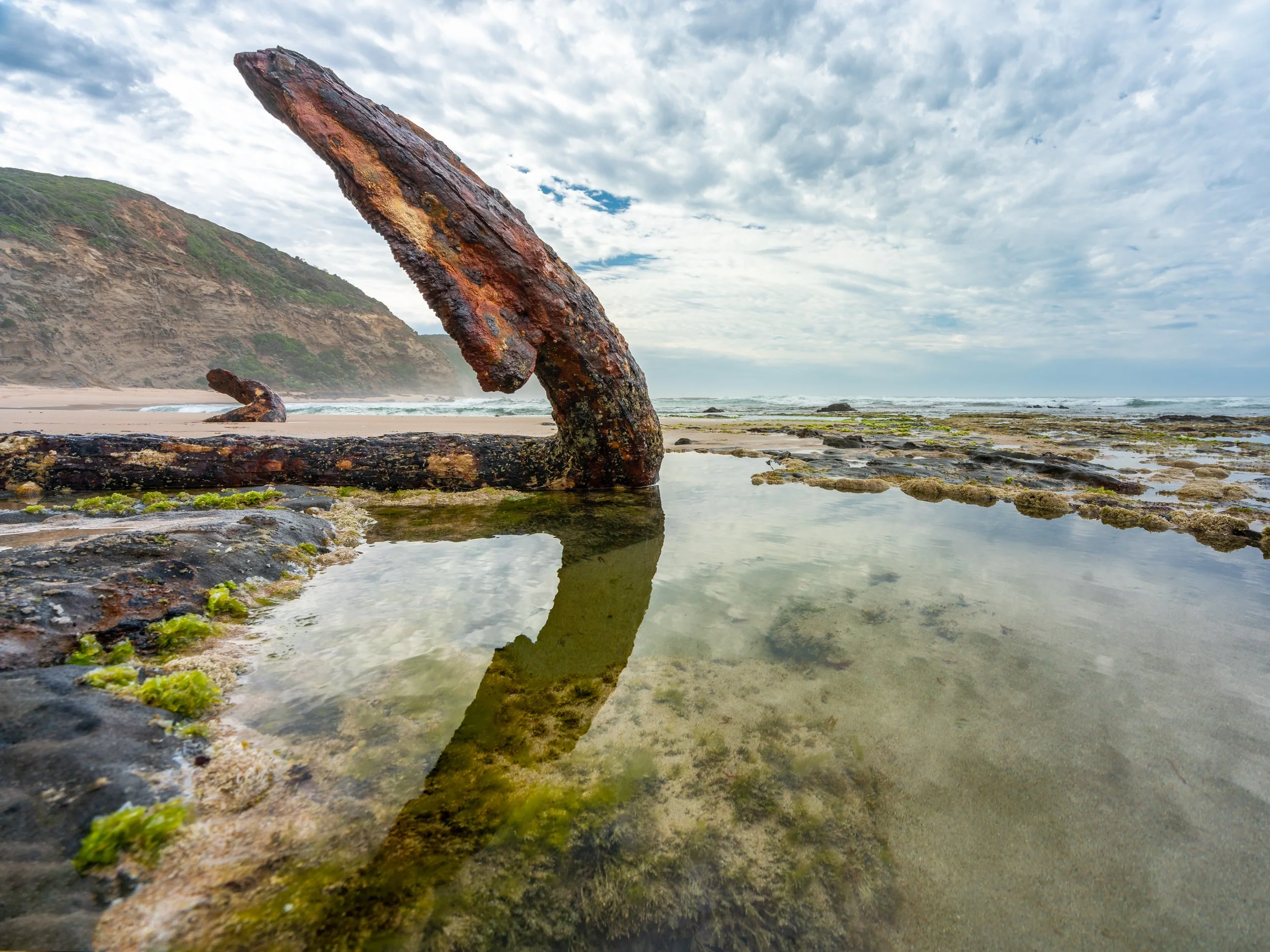 Rusty shipwreck on the beach with tide pools and rocky shoreline, cloudy sky, and a distant hillside in the background.