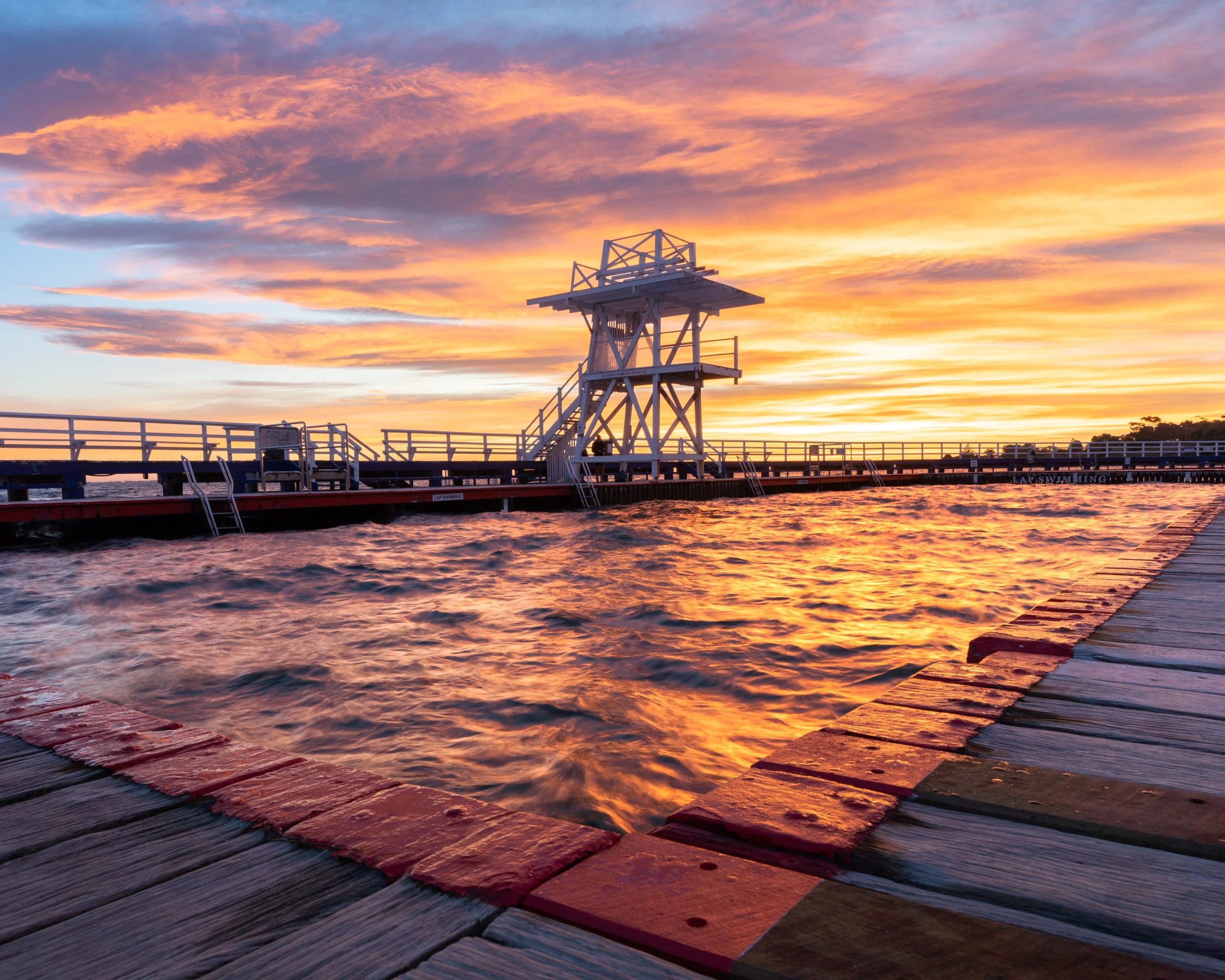 A wooden pier extending into water during a colorful sunset with clouds, featuring a small lifeguard tower.