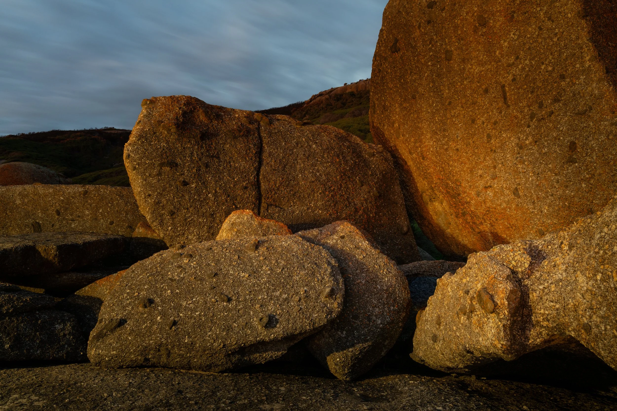 Large rocks and boulders on rocky terrain with a cloudy sky in the background, illuminated by warm sunlight.