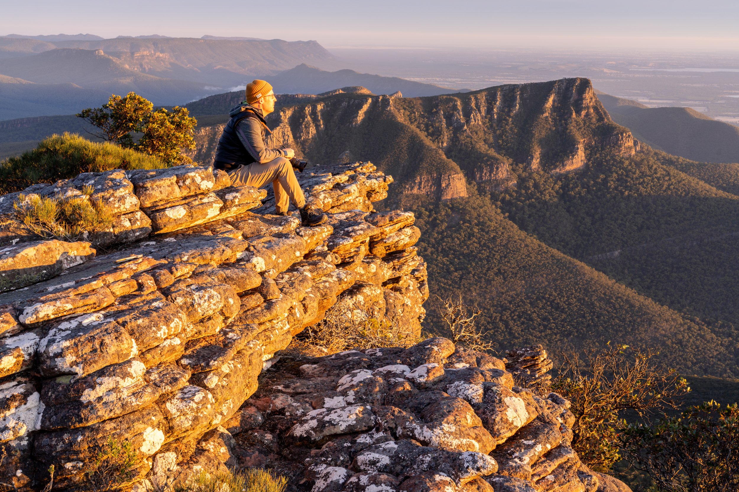 A person sitting on rocky ledge overlooking mountain range at sunset.