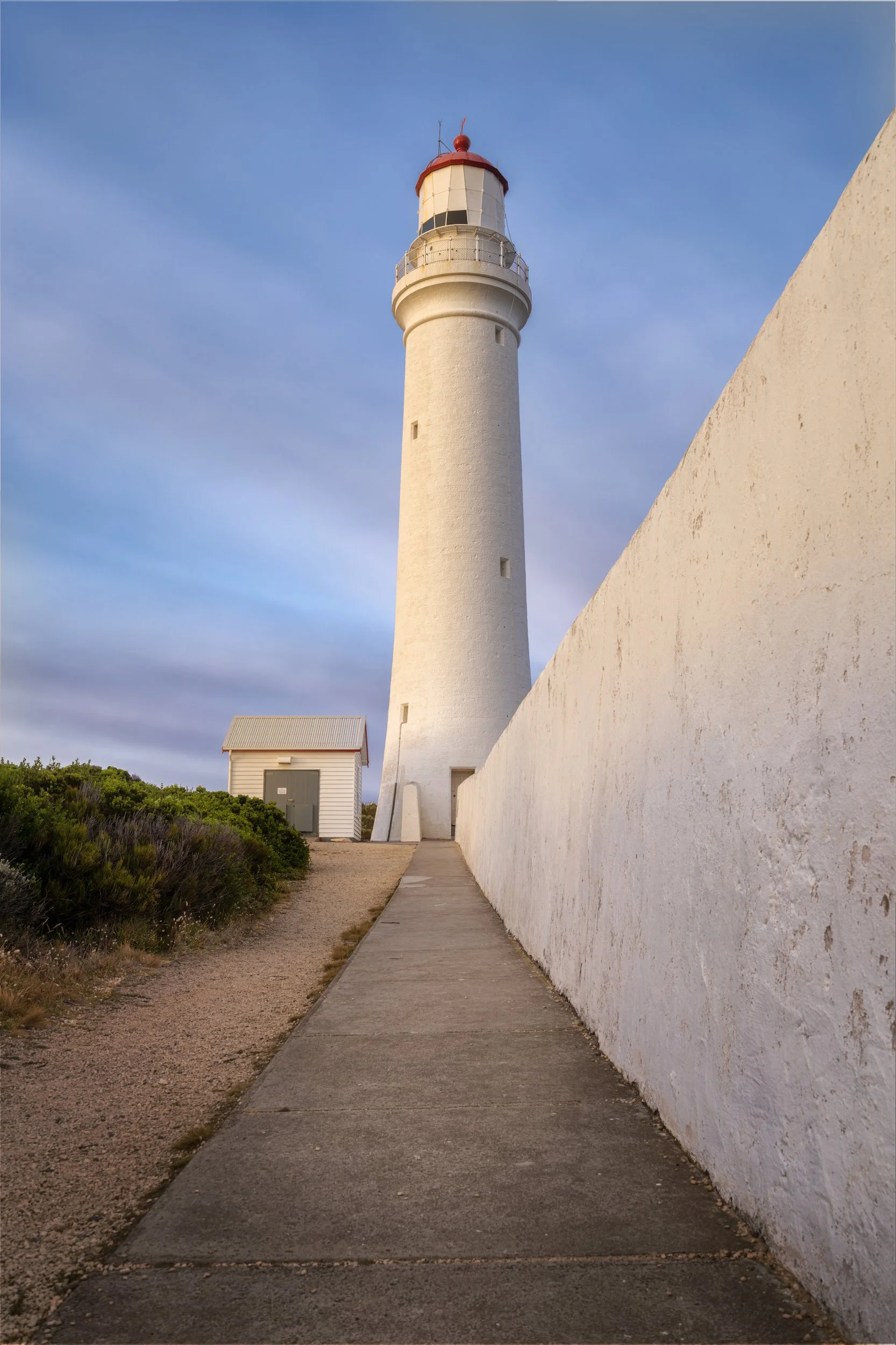 White lighthouse with a red top next to a white wall and a small building, with a cloudy sky background.