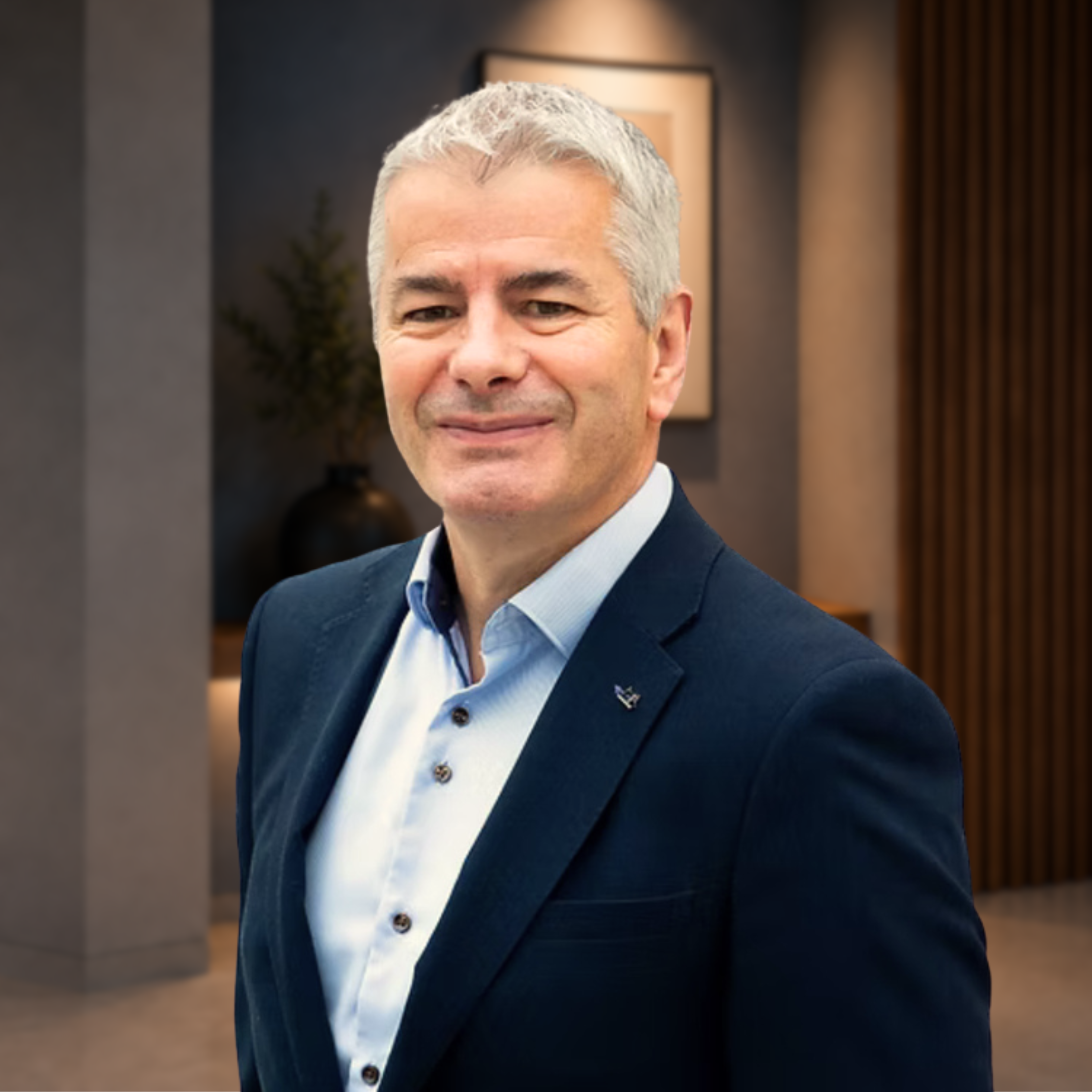 Portrait of a middle-aged man with gray hair wearing a dark suit and light blue dress shirt, smiling against a plain white background.