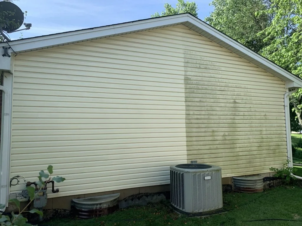 Side view of a house with vinyl siding, partially cleaned and dirtier area on the right, an air conditioning unit on the ground, and trees in the background.