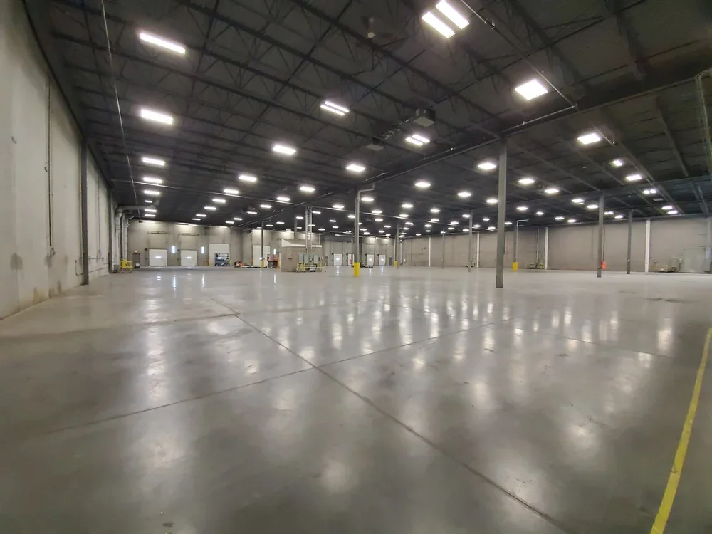 Empty warehouse with polished concrete floor and high ceiling, illuminated by overhead lights, with several doors and equipment at the far end.