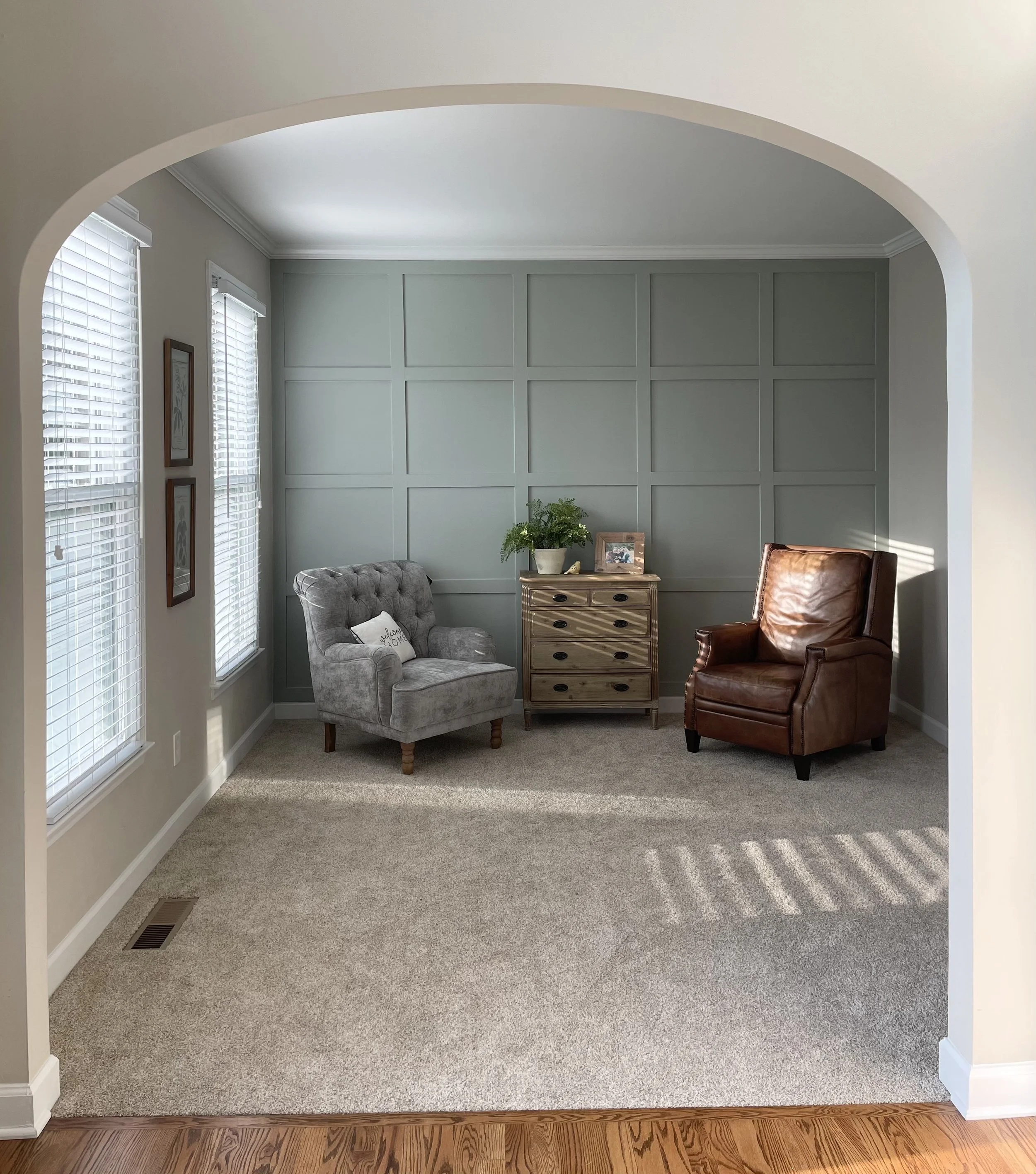 A cozy sitting area with gray and brown armchairs, a wooden side table with a plant, picture frame, and a small decorative object, illuminated by sunlight coming through two windows with white blinds, in a room with light-colored walls and carpeted floor.