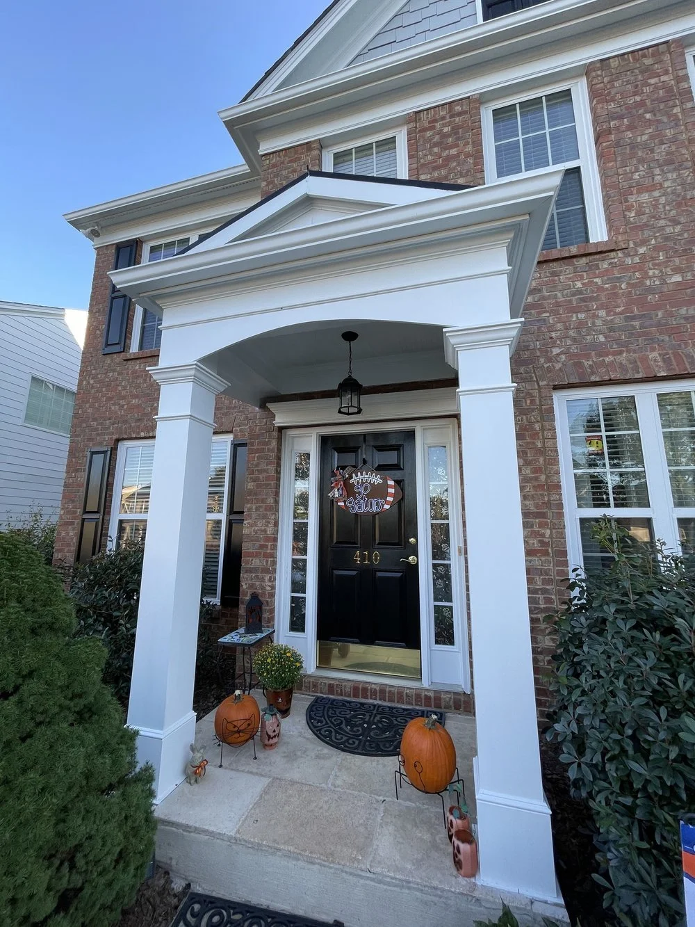 Front door of a brick house decorated for fall, with pumpkin planters, gator wreath, and holiday signs, under a white porch.