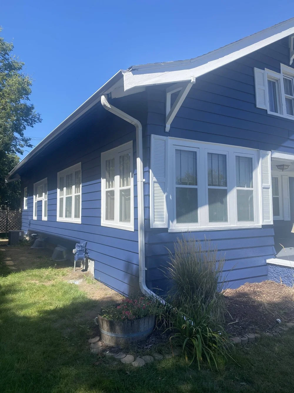 A blue house with white trim, multiple windows, a manicured lawn, and a flower bed in a circular wooden container under bright sunlight.