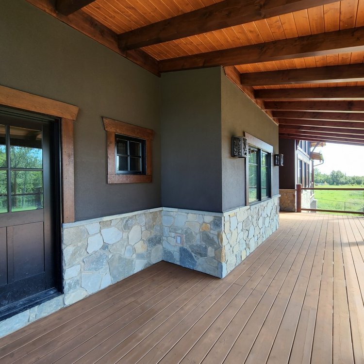 Exterior view of a house porch with wooden flooring, stone and dark gray wall, and a wooden ceiling. There are large windows and a door, with trees and a field in the background.