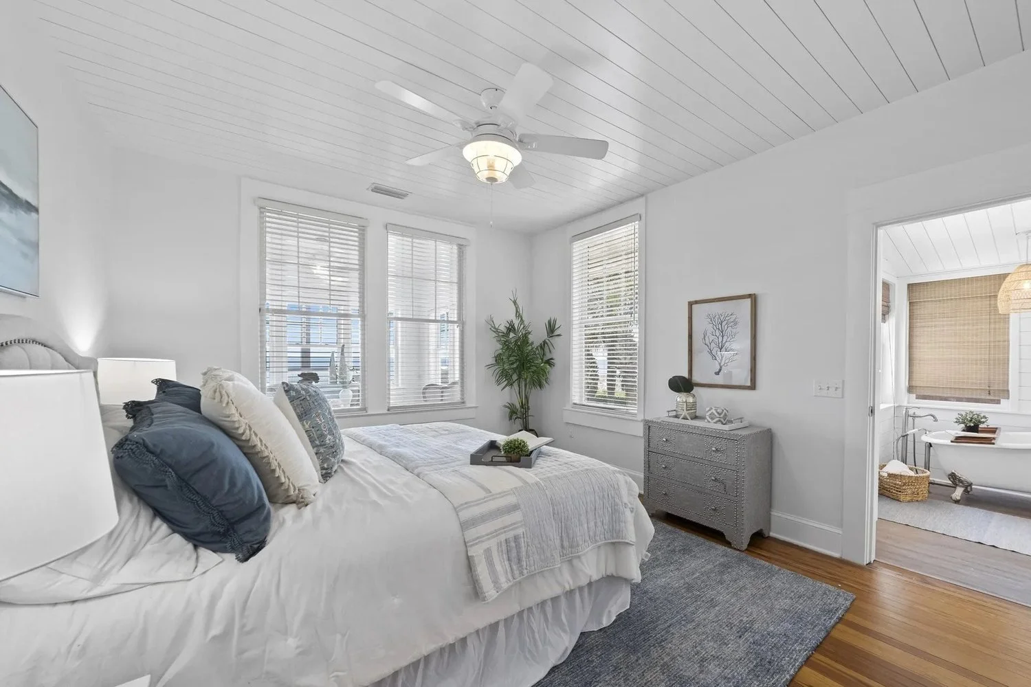 A bright, white bedroom with a bed, pillows, and a tray on top, alongside a green plant, and a chest of drawers decorated with artwork, in a room with wooden floors, windows with blinds, and an adjoining bathroom with a bathtub and a window with a ba