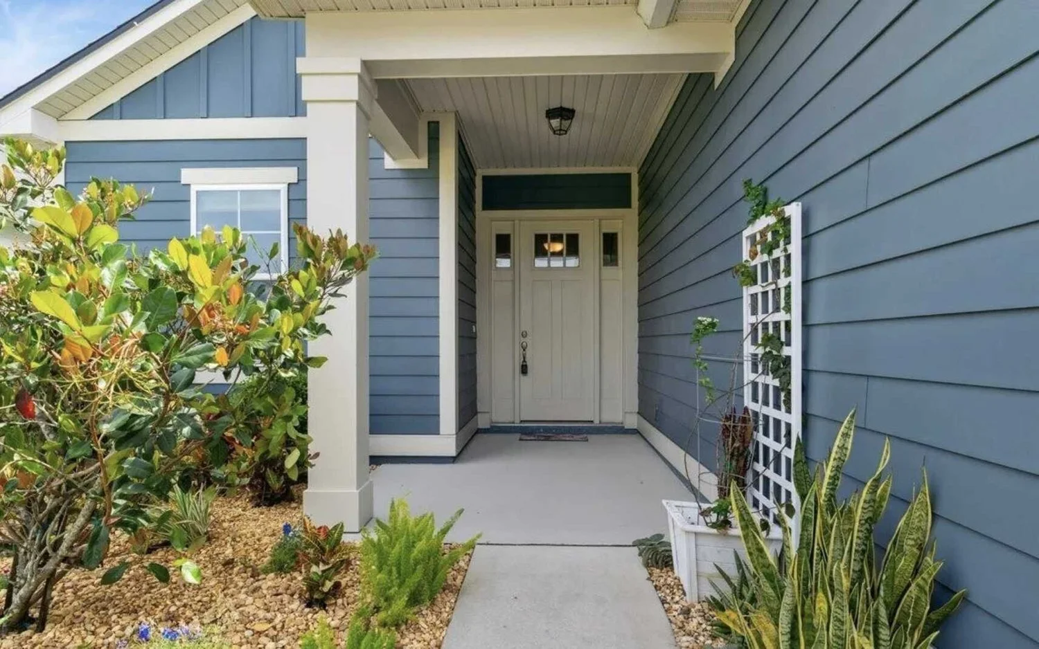 Front porch of a house with blue siding, white trim, a white door, and a small window above it, surrounded by plants and landscaping.