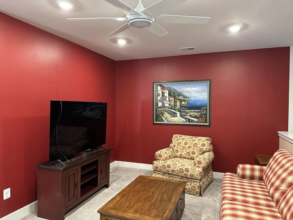 Living room with red walls, a flat-screen TV on a wooden stand, a floral armchair, a checkered sofa, a wooden coffee table, a framed artwork of a coastal scene, a ceiling fan, and recessed lighting.