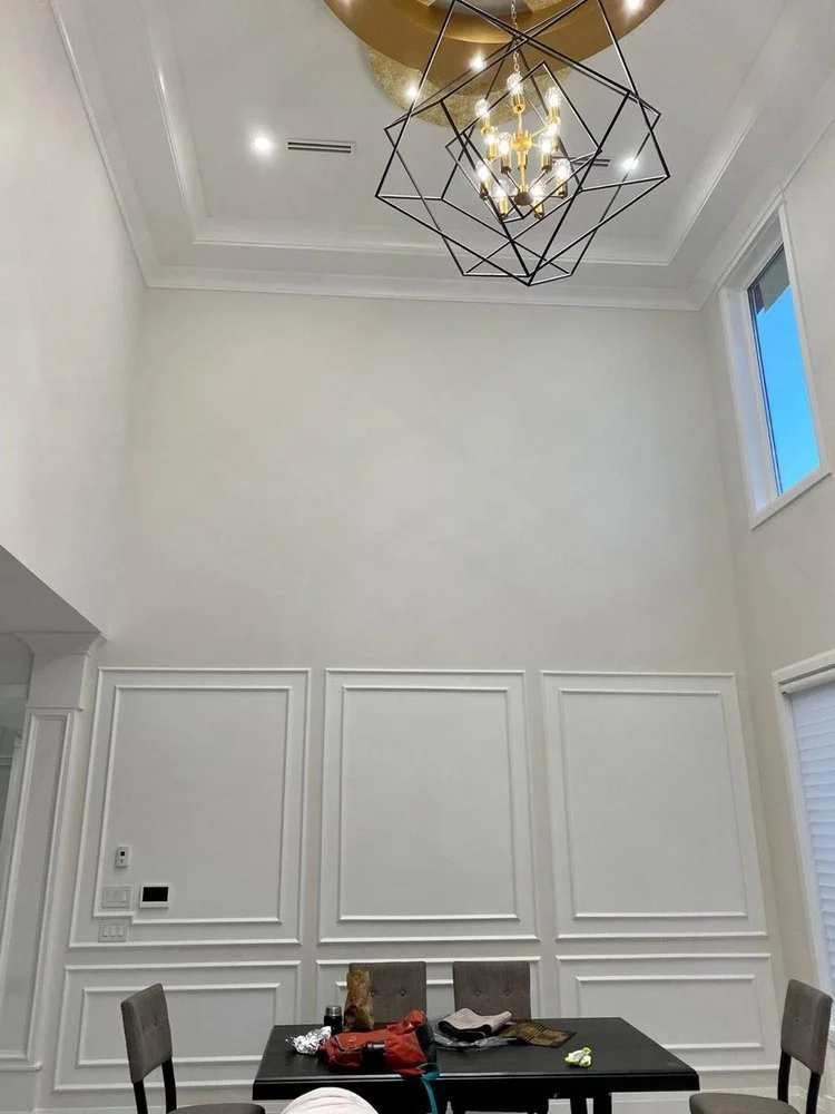 Interior room with high ceiling, modern geometric chandelier, white paneled walls, small window, and a dining table with chairs, items on table including a red bag, hat, and other objects.