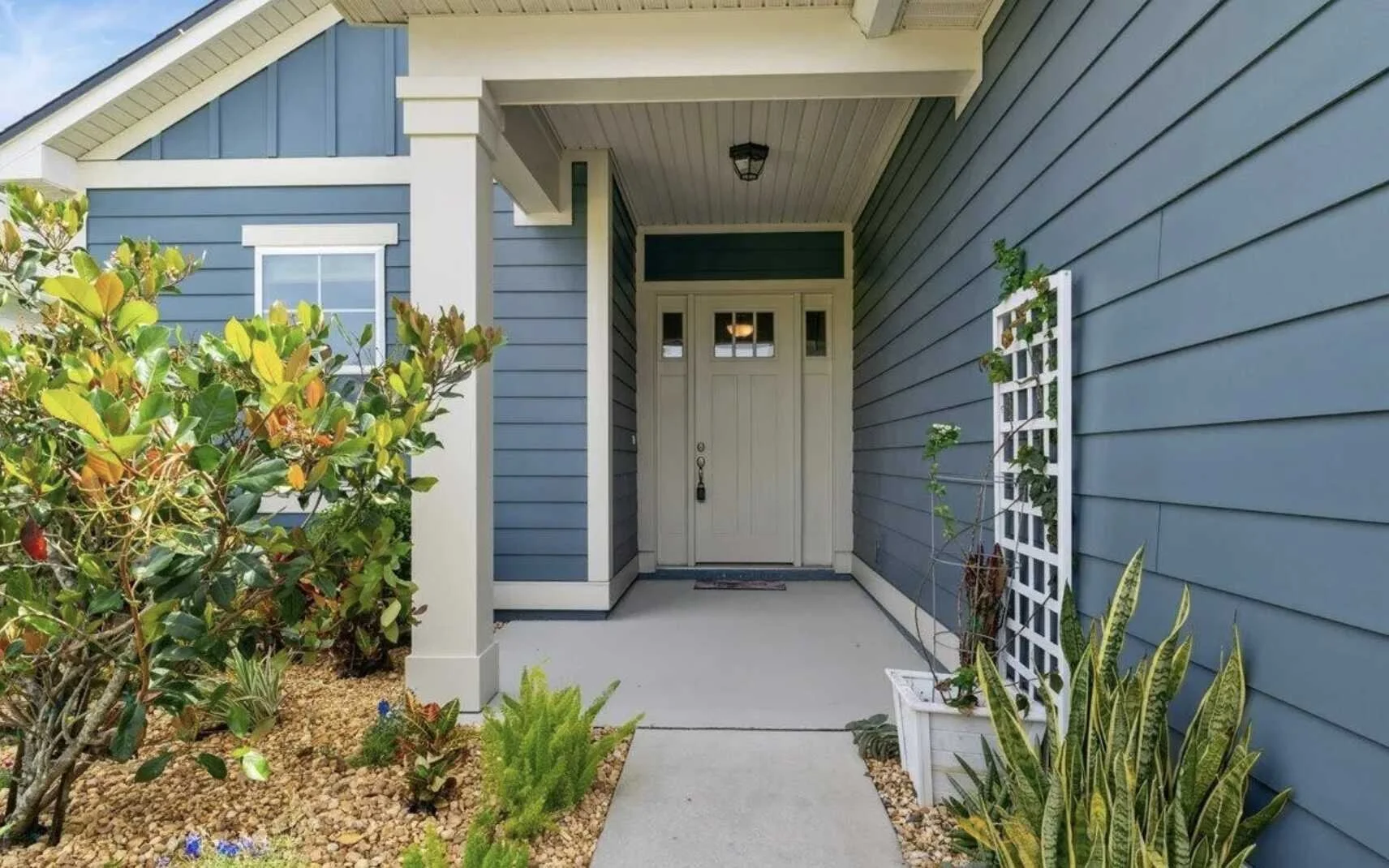 Front porch of a house with blue horizontal siding and a white door, surrounded by potted plants and greenery.