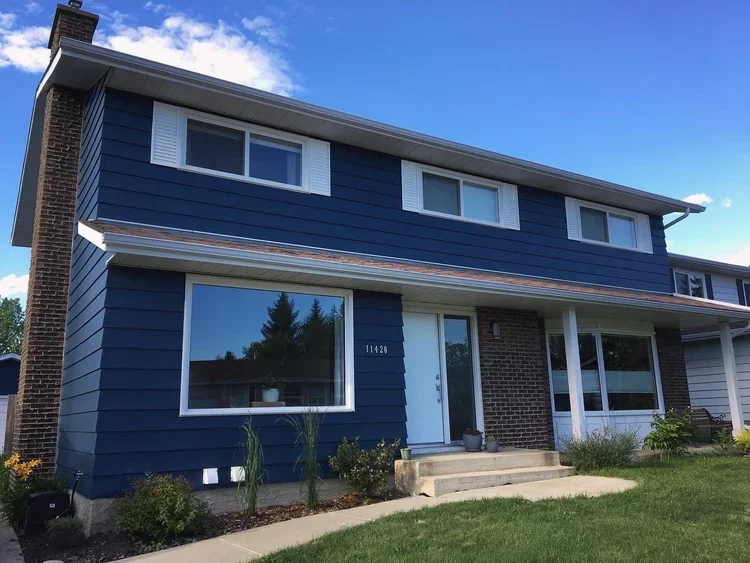 A two-story house with blue siding, white trim, and a brick accent on the front wall. There are four windows on the second floor and two large windows on the first floor, with a white front door and steps leading up to it. The house has a front lawn 
