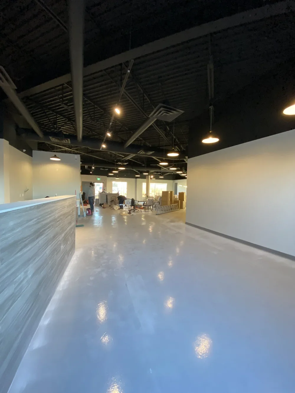 Interior of a room under construction with a shiny polished floor, a reception counter on the left, and workers assembling furniture in the background, illuminated by hanging ceiling lights.